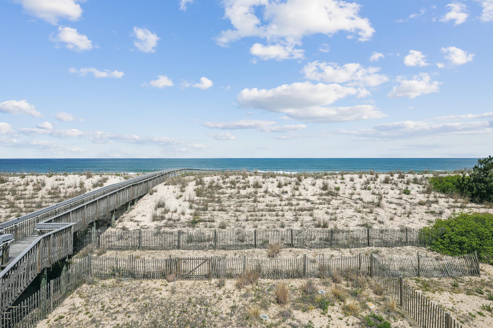 The property features a wooden beach access walkway leading to the sandy shoreline, with protected dune vegetation and an expansive view of the Atlantic Ocean under a bright, partly cloudy sky.