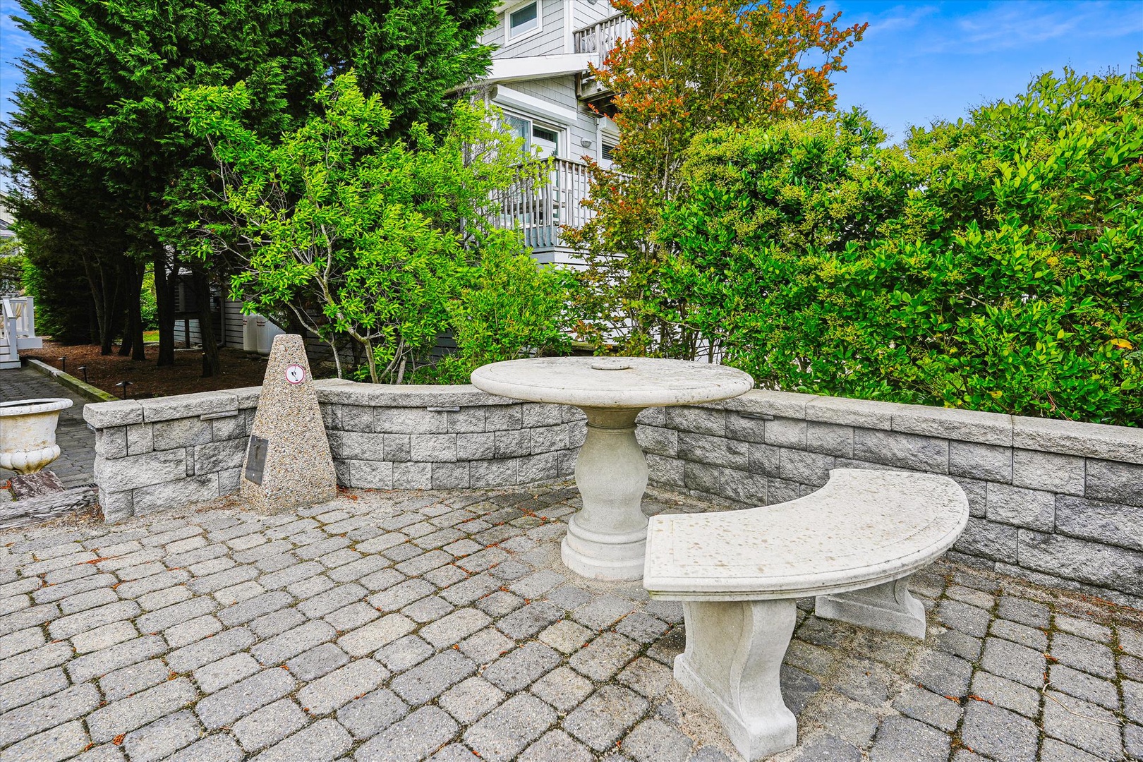 Paver stone courtyard with decorative fountain centerpiece, framed by lush greenery and mature trees near the property's white-sided building.