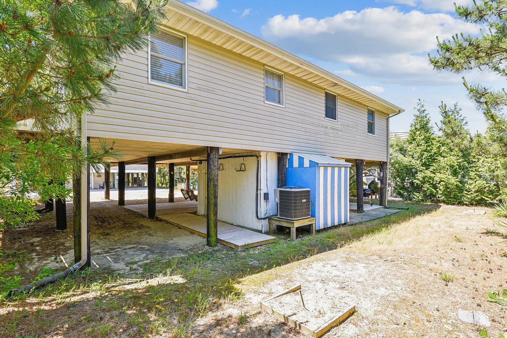 Coastal-style vacation home features covered parking and outdoor storage beneath the main living area, surrounded by mature pines and greenery.