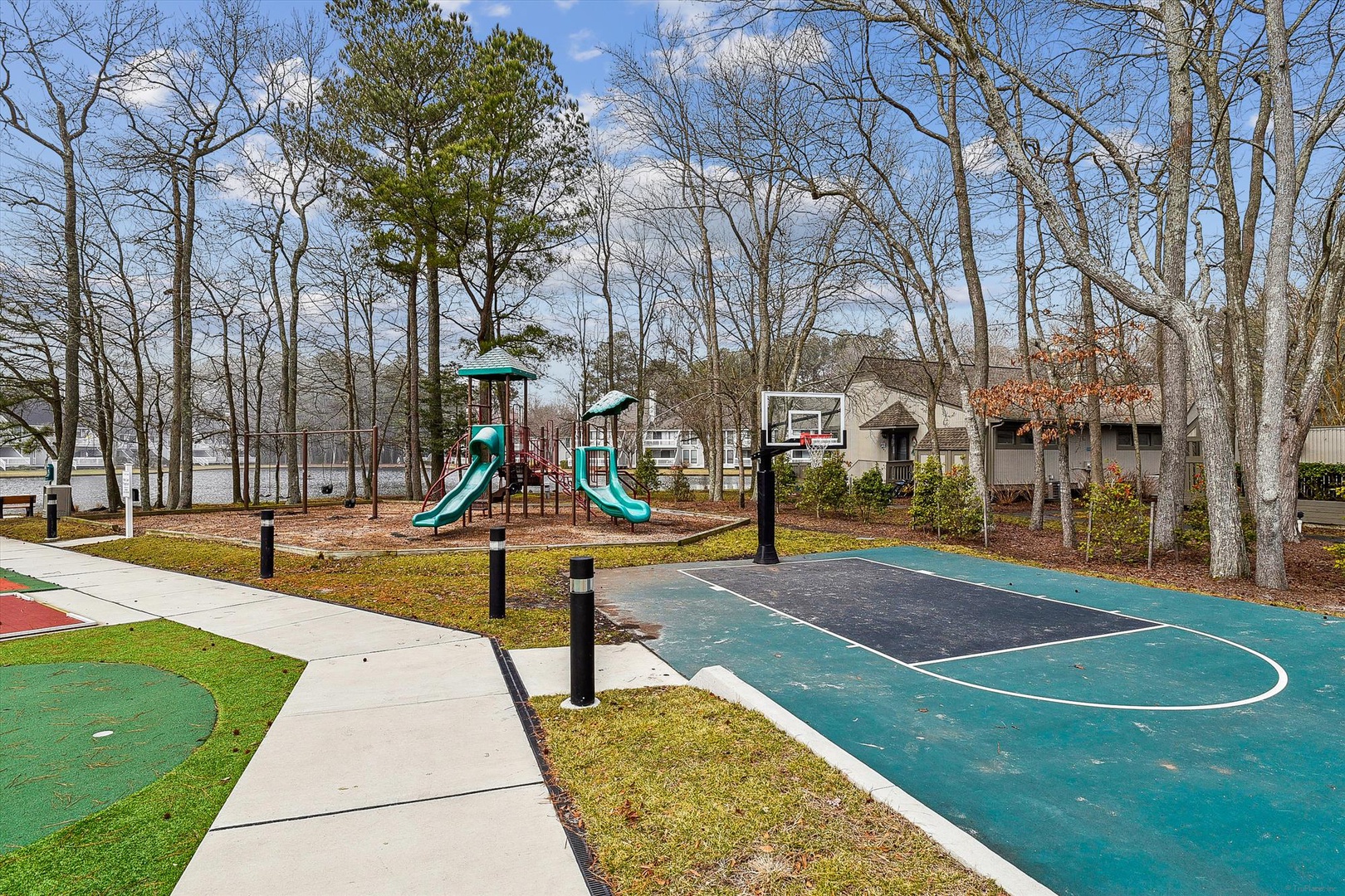 Lakeside community featuring a playground with slides, outdoor basketball court, and walking paths among tall trees, with homes visible in the background.