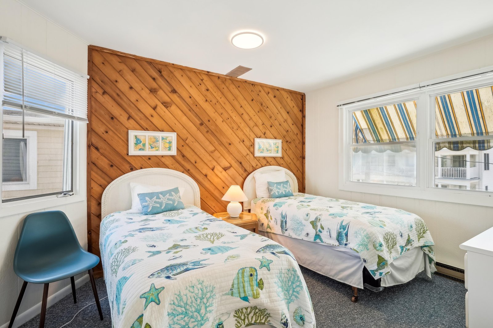 Two cozy twin beds await you in this coastal-themed bedroom, featuring cheerful ocean-print bedding and a warm wood accent wall that adds rustic charm to your beach escape.