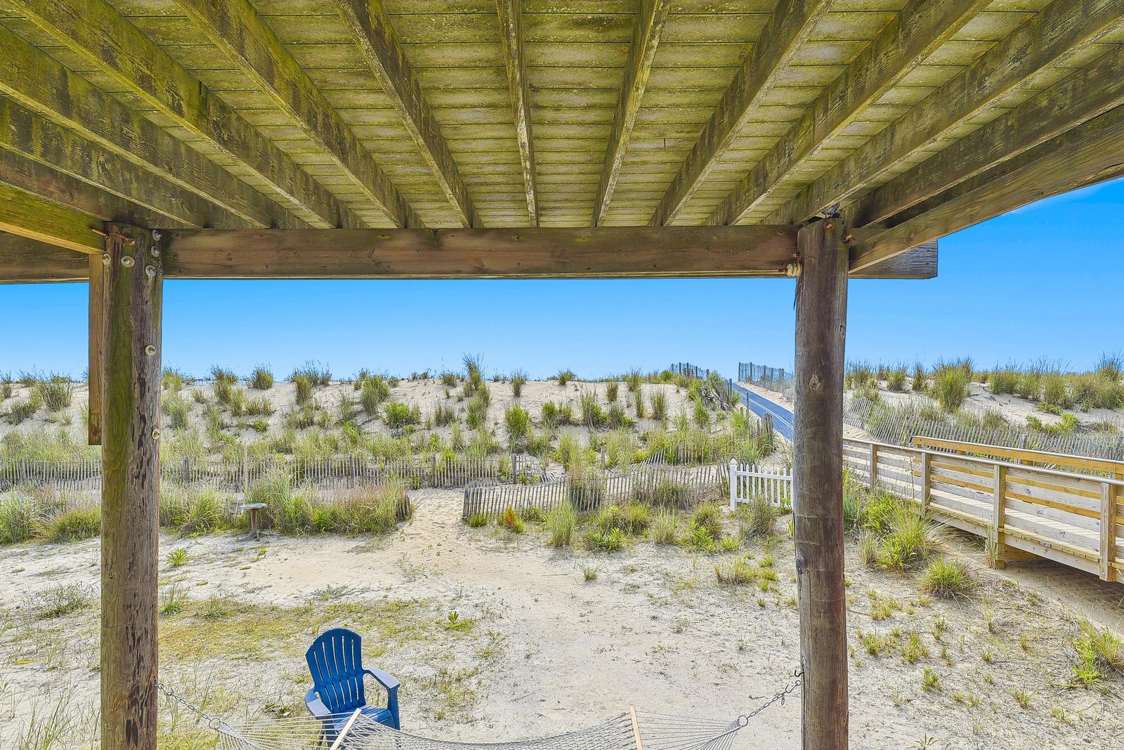 Relax in your beach chair beneath the covered deck while soft sand, dune grasses, and open blue sky stretch before you—your quiet coastal escape.