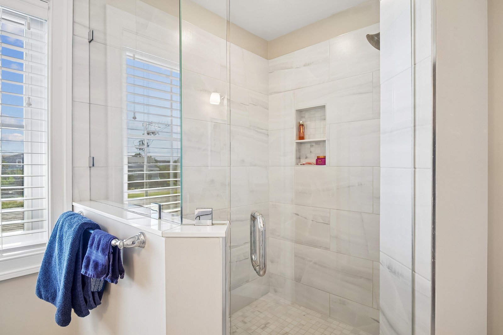 Step into your elegant walk-in shower with marble-style tile, a built-in niche for your essentials, and natural light from the window to brighten your morning routine.