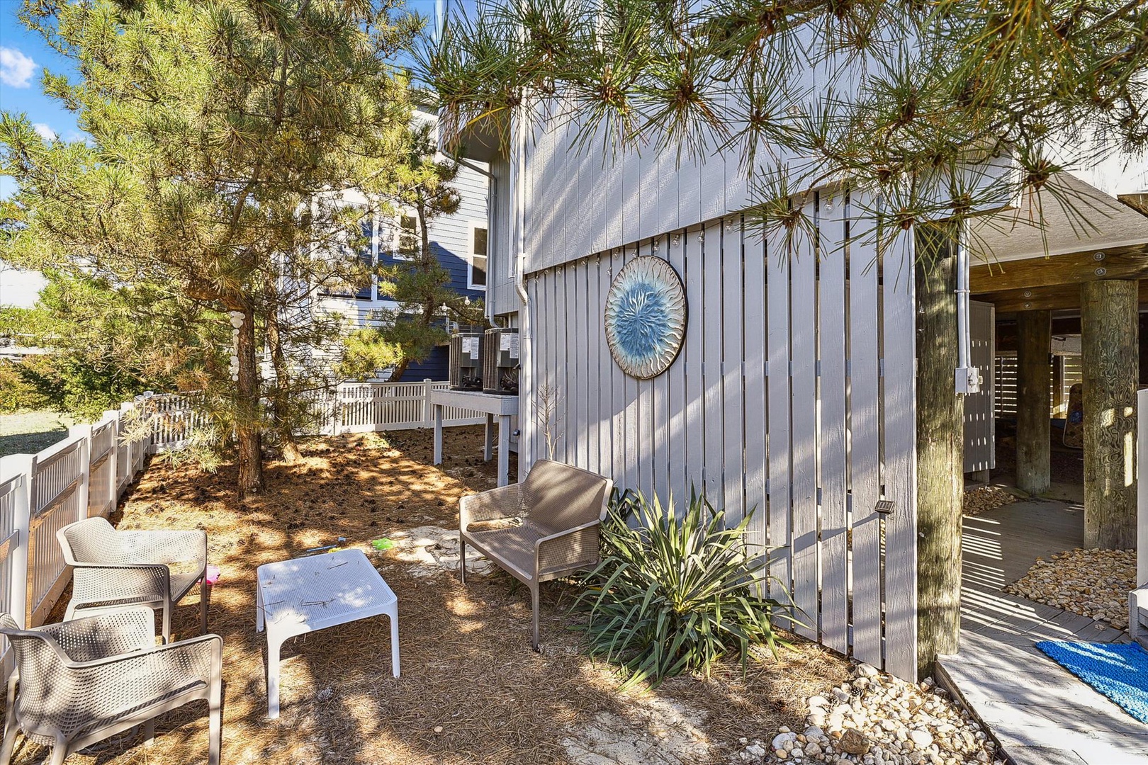 This private outdoor courtyard features shaded seating beneath tall pines, offering a peaceful retreat for morning coffee or evening relaxation. The decorative wall art and natural landscape create a calm coastal atmosphere.