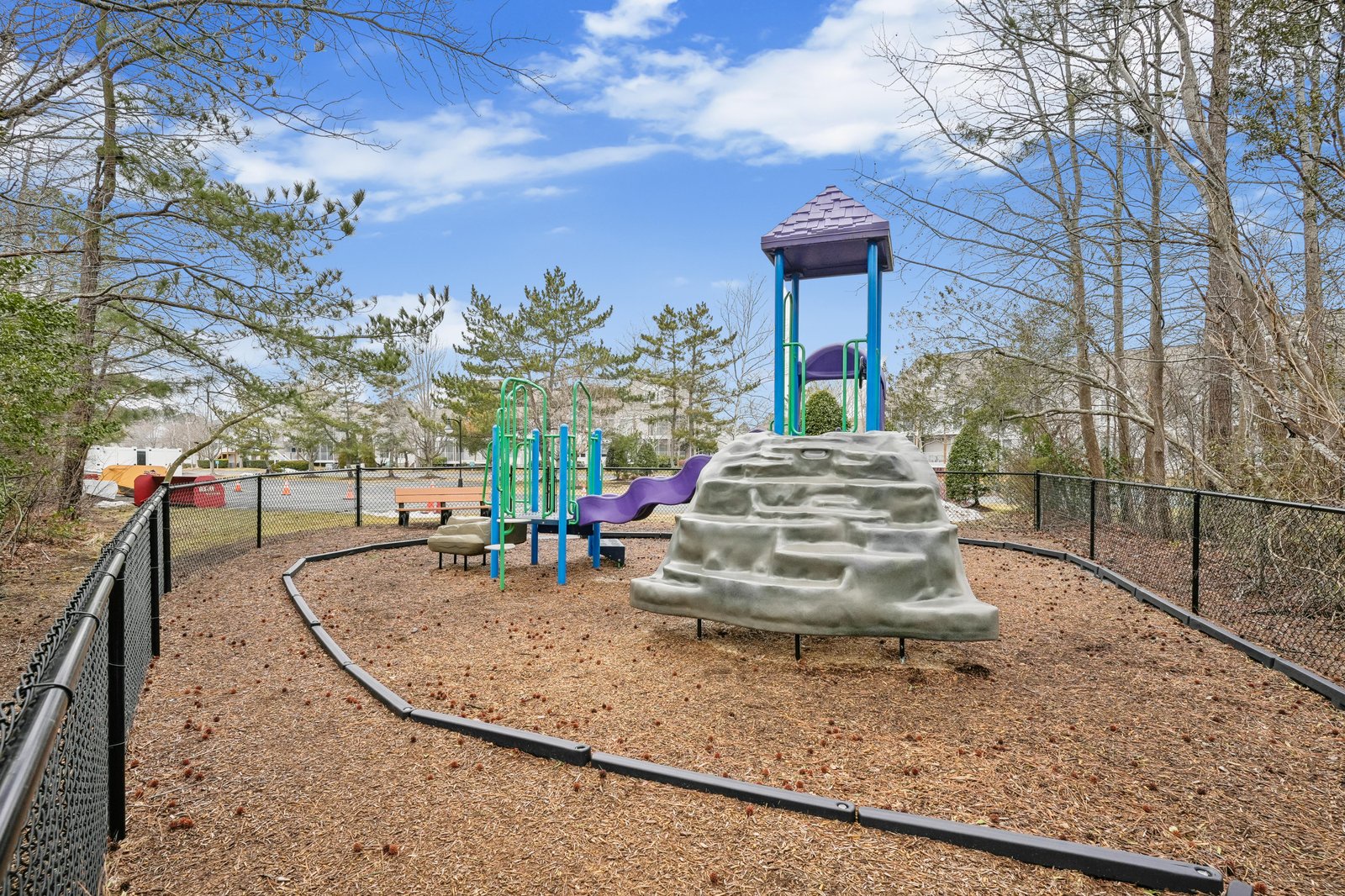 A fenced playground features climbing structures and slides surrounded by towering pines, with parking and buildings visible beyond the play area.