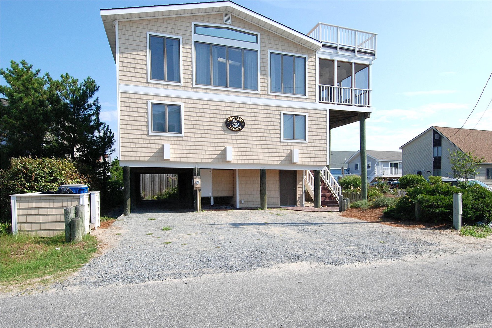 Coastal vacation home with elevated stilts, dual-level decks, and gravel parking. The property features modern siding, large windows, and a convenient covered ground-level area near neighboring beach houses.