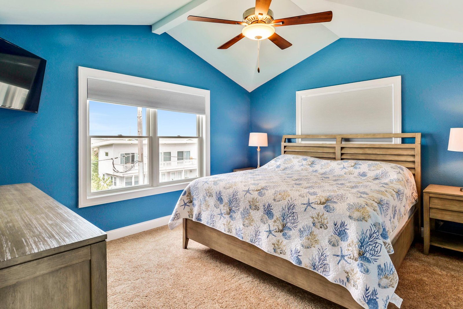Your serene coastal-themed bedroom features vaulted ceilings, a ceiling fan, and a bright window framing nearby buildings. Crisp blue walls and beach-inspired bedding create a peaceful retreat.