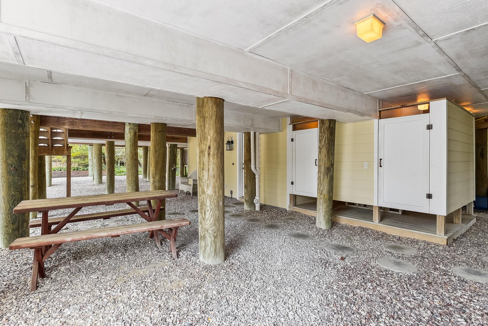A covered outdoor area beneath the property features rustic log posts, gravel flooring, and natural light filtering through. A picnic table invites you to relax in this sheltered, wood-accented space.