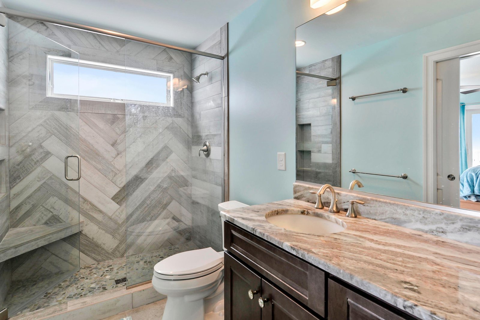 Step into your private spa-like retreat with a stunning walk-in shower featuring designer tile in a striking chevron pattern, elegant marble countertops, and soft coastal blue walls that set a serene, calming tone.