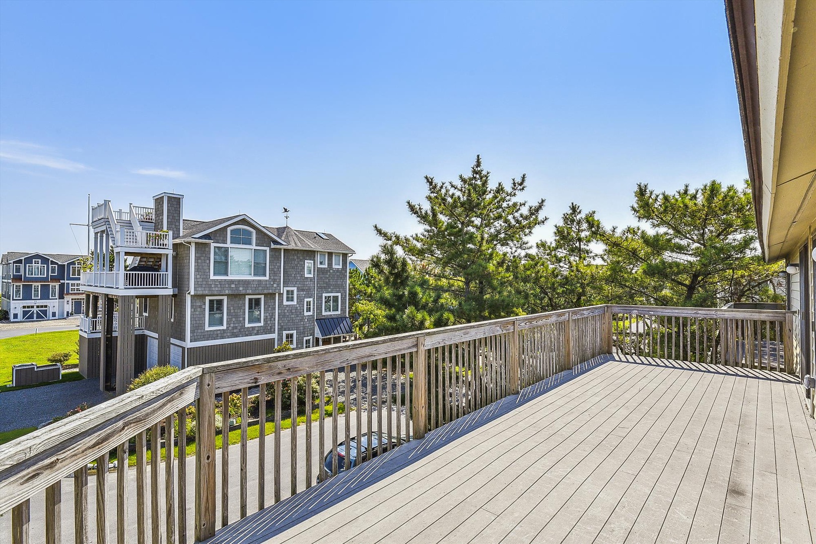 Spacious deck overlooking coastal trees and neighboring beach homes, with plenty of room for outdoor lounging under clear blue skies.