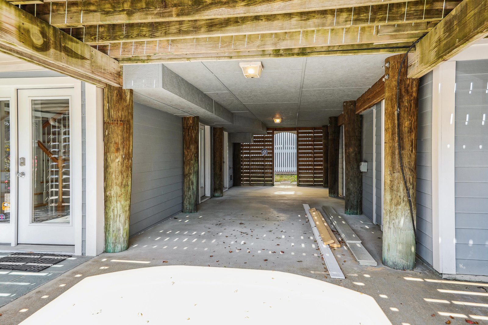 A covered entry area beneath the home features rustic timber posts and opens to a dappled sunlit outdoor space, framing your arrival with coastal character and natural charm.