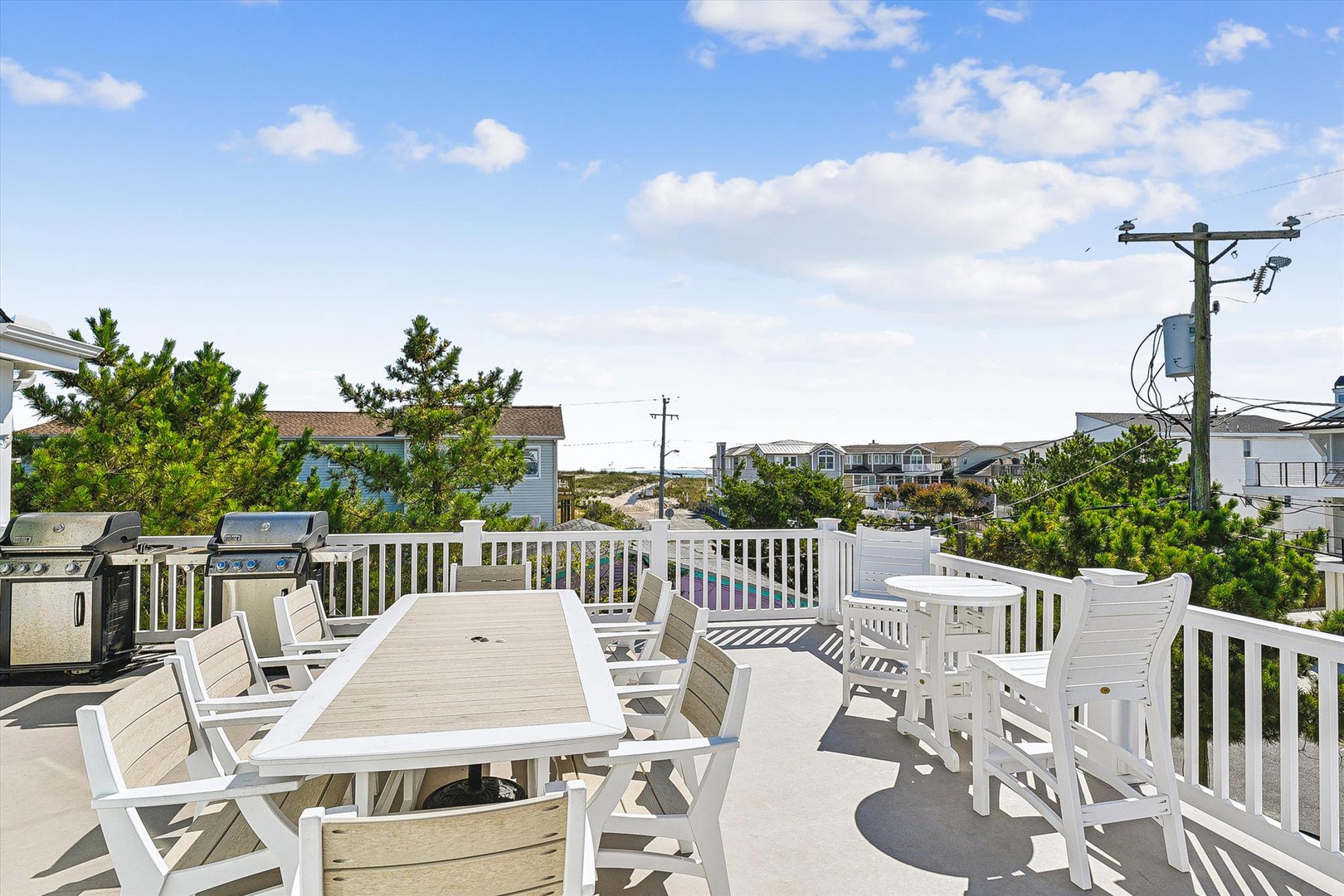 A spacious rooftop deck with dining and lounge areas awaits your morning coffee or evening gatherings. Enjoy coastal breezes, comfortable seating, and grilling space under open skies, surrounded by glimpses of nearby homes and greenery.