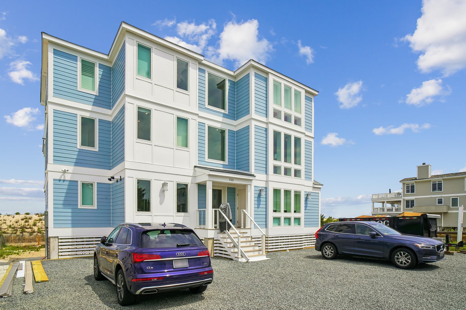 Modern three-story beachside rental featuring fresh coastal-blue siding and a spacious gravel parking area, situated in a quiet seaside neighborhood.