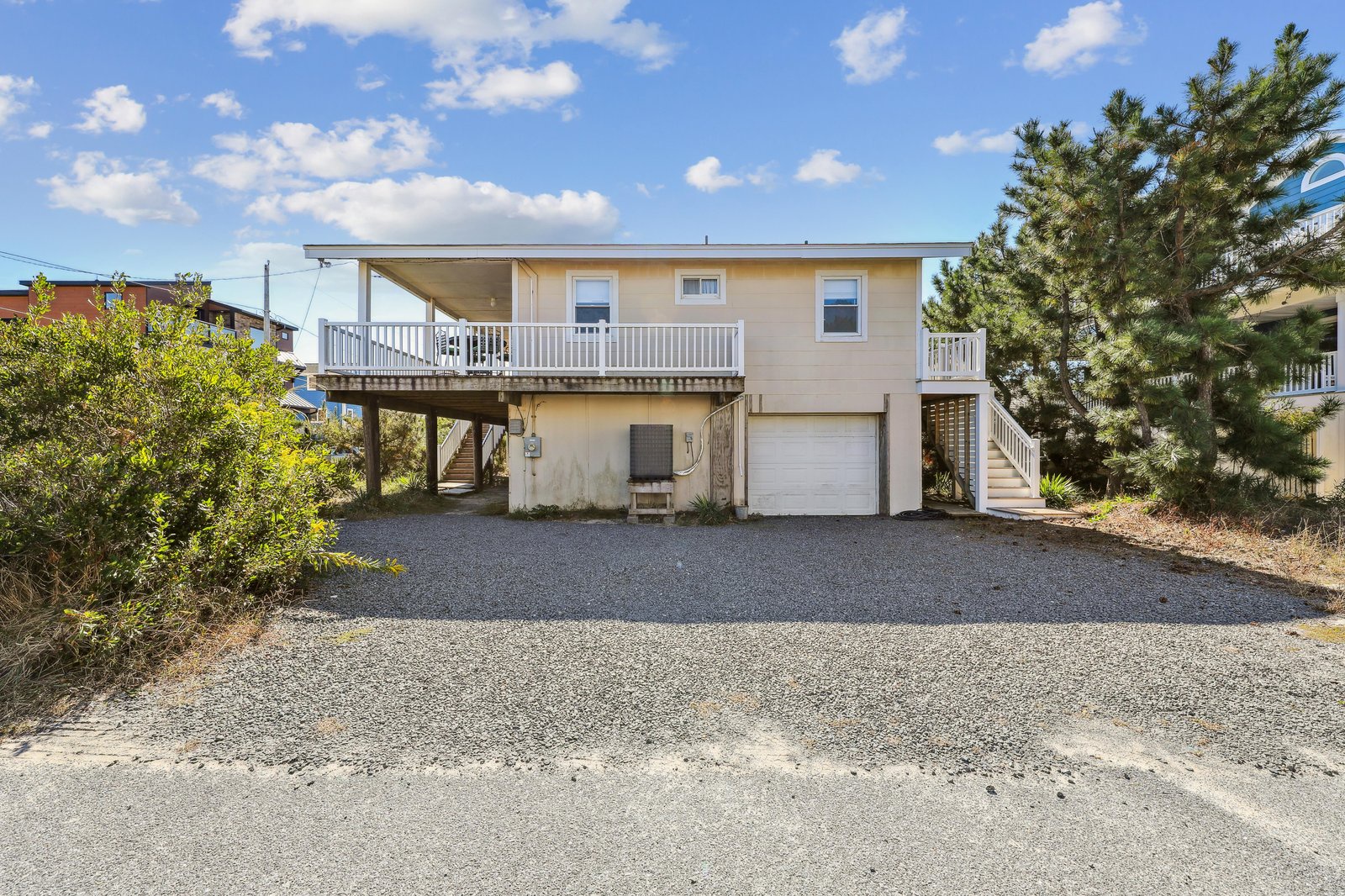 The property features a two-story beachside structure with a spacious upper-level deck and convenient parking below. The home is nestled among coastal greenery and offers easy access to the beach.