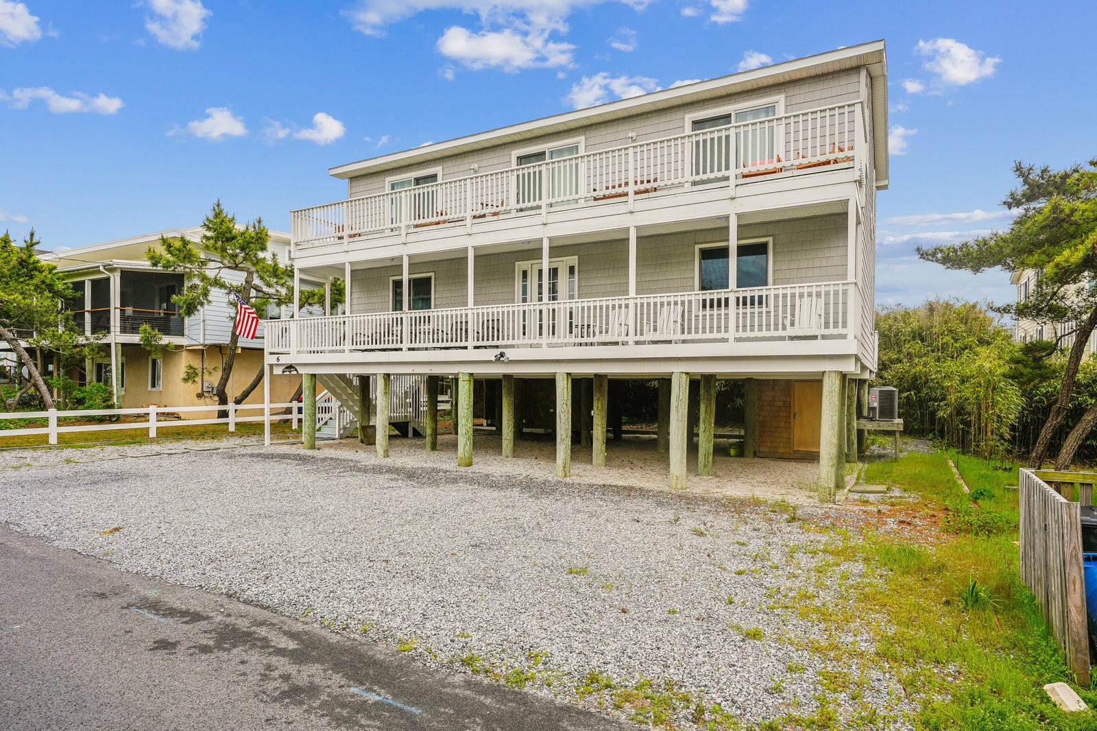 A spacious coastal rental elevated on pilings with ample parking and multiple balconies surrounded by lush greenery beneath a bright blue sky.