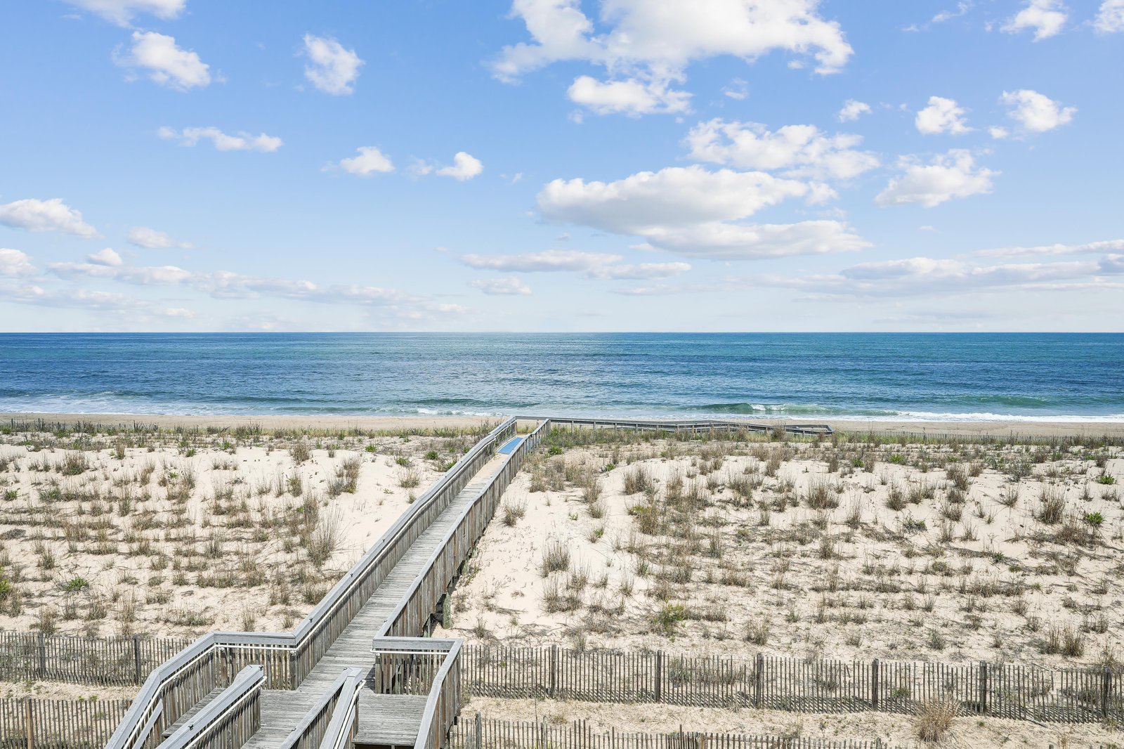 A wooden boardwalk leads across pristine dunes to the open ocean, offering direct access to a beautiful sandy beach with gentle waves rolling ashore under an expansive blue sky.