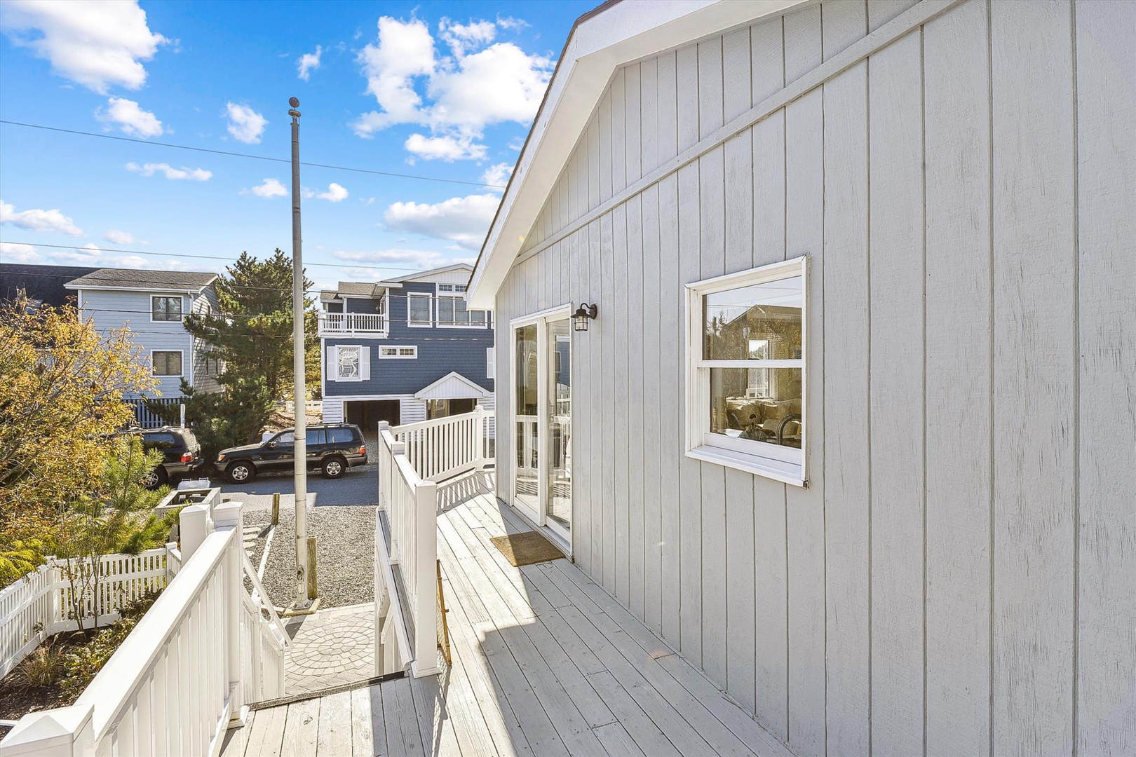 A charming coastal property nestled in a quiet beach neighborhood, featuring bright white architectural details and surrounded by neighboring homes. The exterior showcases classic beach-style siding under a clear blue sky.