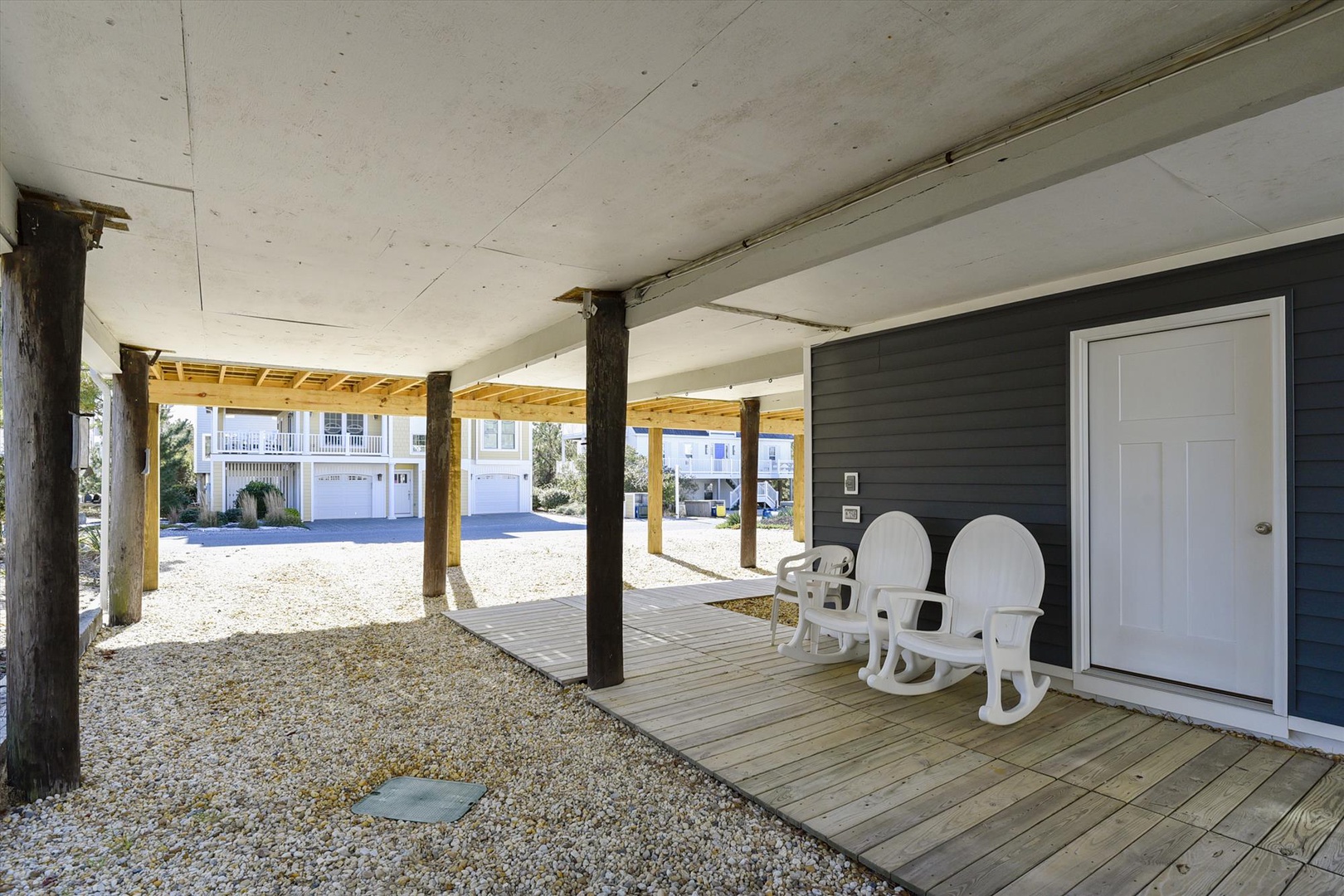 Covered ground-level parking area beneath a raised beach house with wooden deck seating, gravel drive, and coastal neighborhood views beyond.