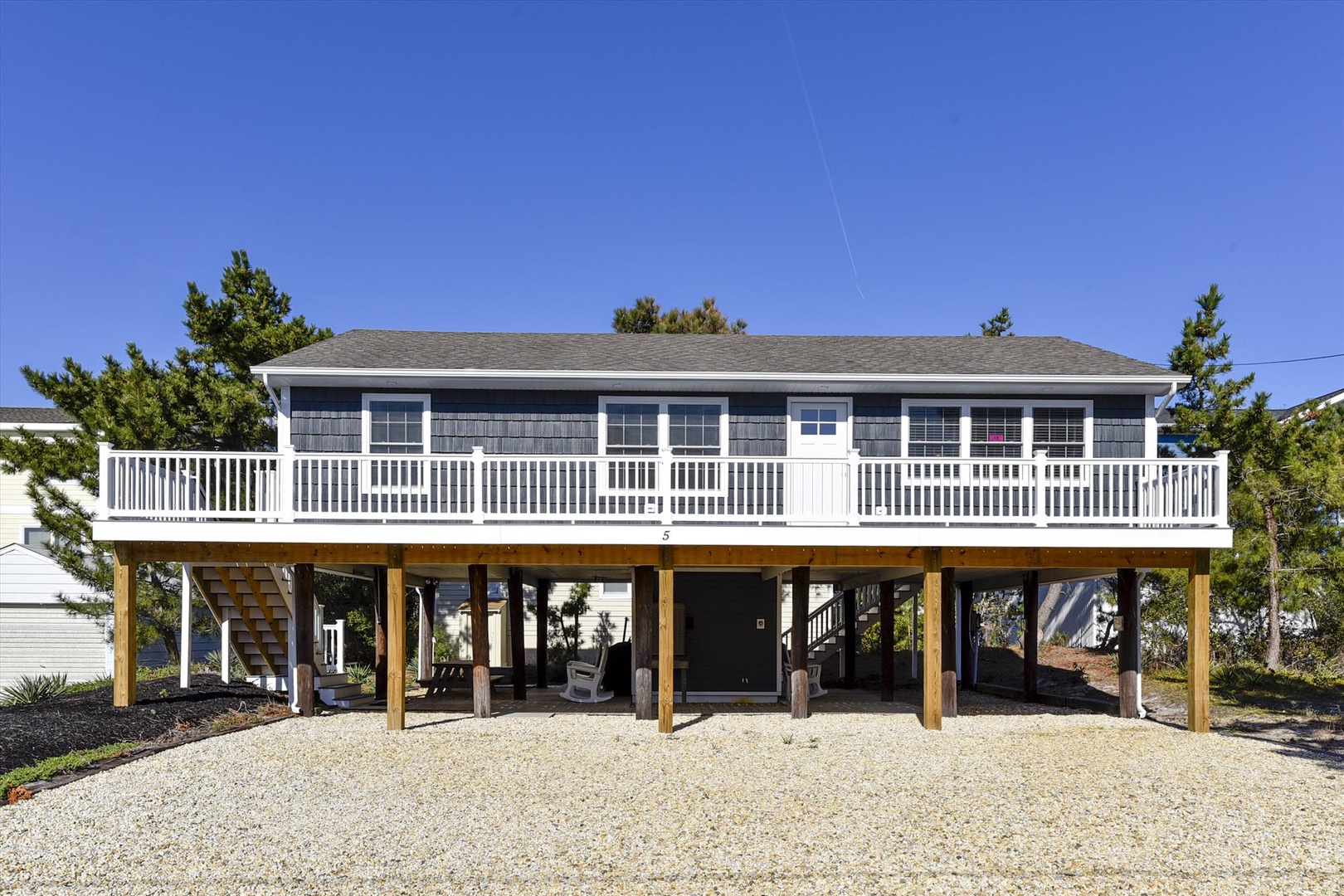 Charming coastal cottage elevated on stilts with wraparound deck and gravel parking area surrounded by mature trees.