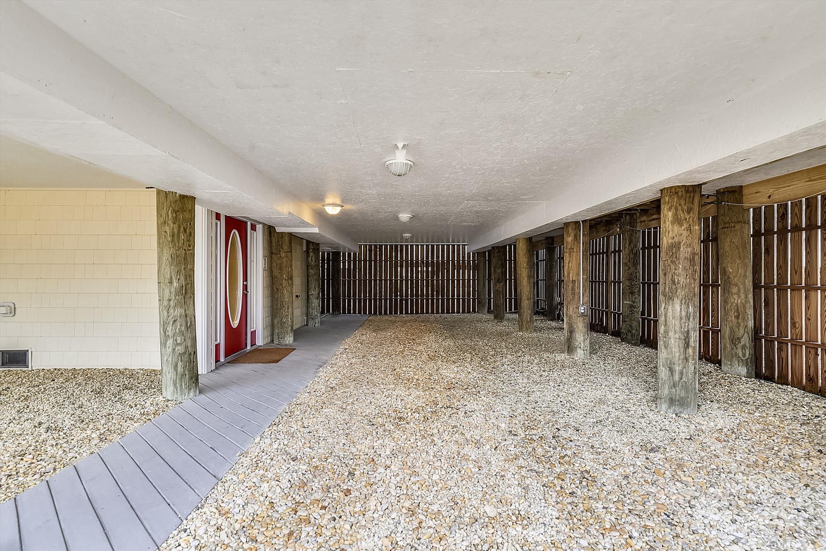 Covered ground-level parking with gravel flooring, rustic wooden support beams, and direct access to the property entrance through the distinctive red-accented door.