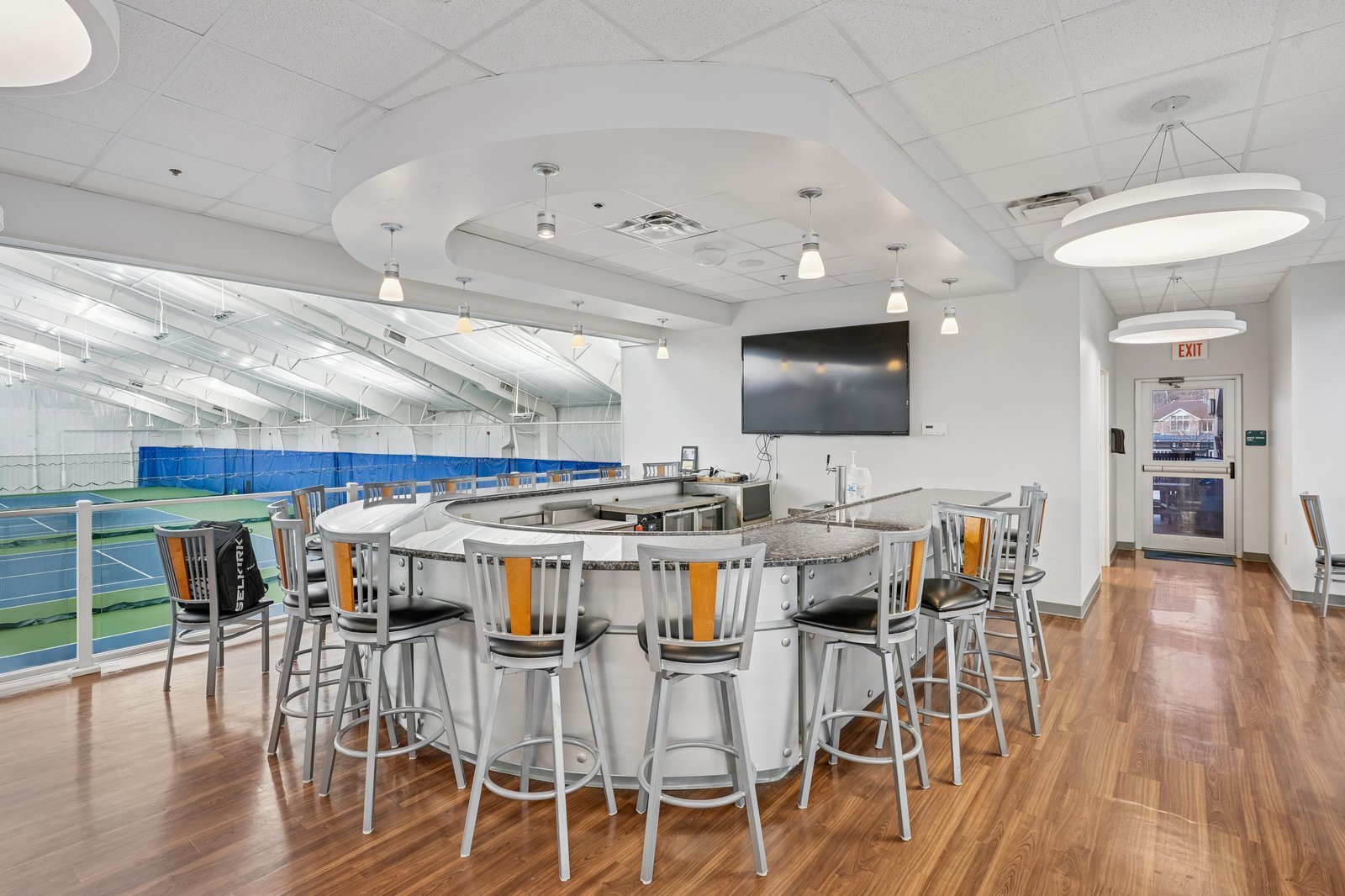 Modern clubhouse lounge overlooking indoor tennis courts, with bar seating, TV, and bright natural light—perfect for relaxing after your match.