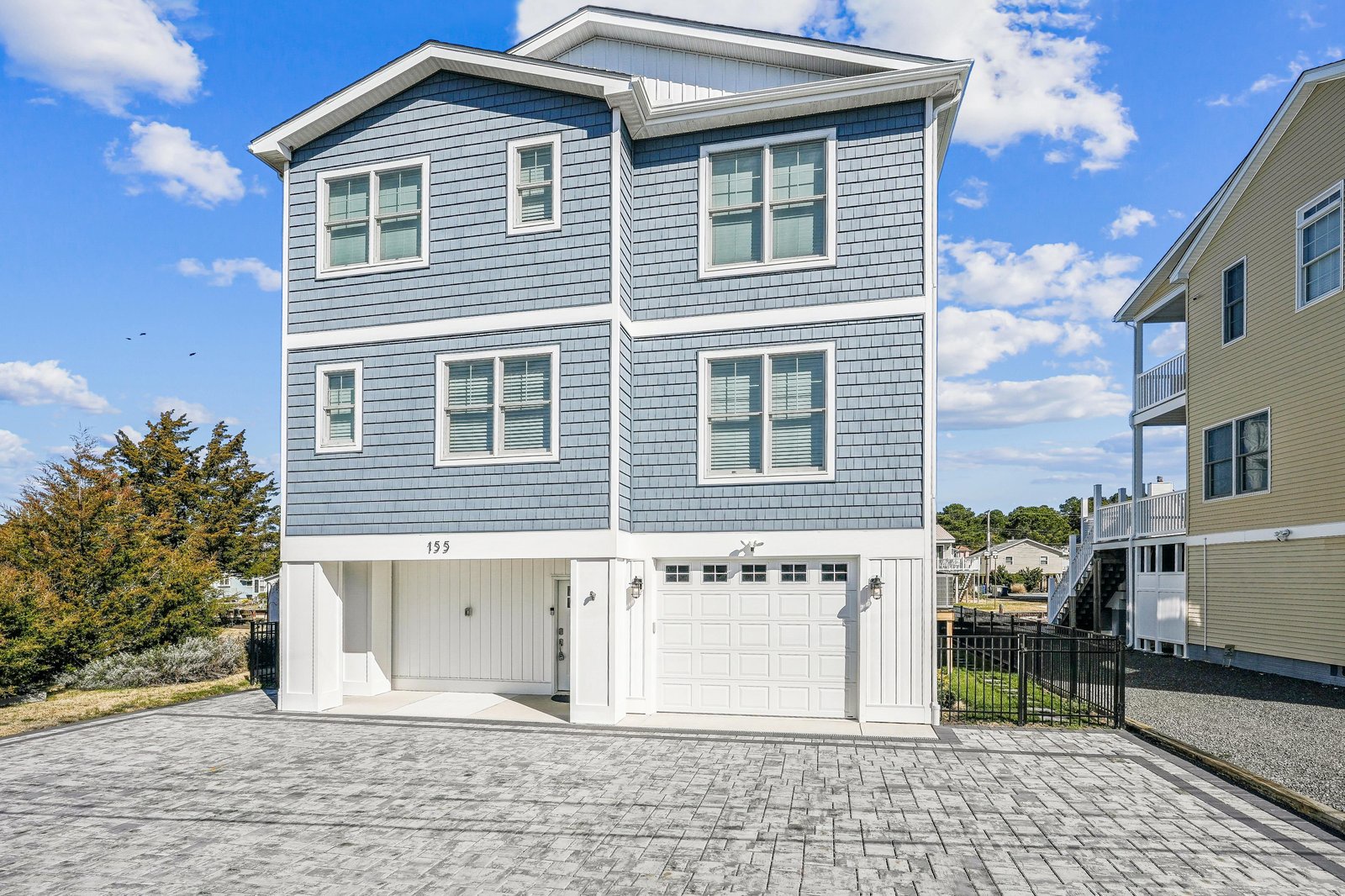 A coastal-style vacation home with charming blue-gray siding and bright white trim, featuring a two-car garage and convenient ground-level parking on a paver driveway.