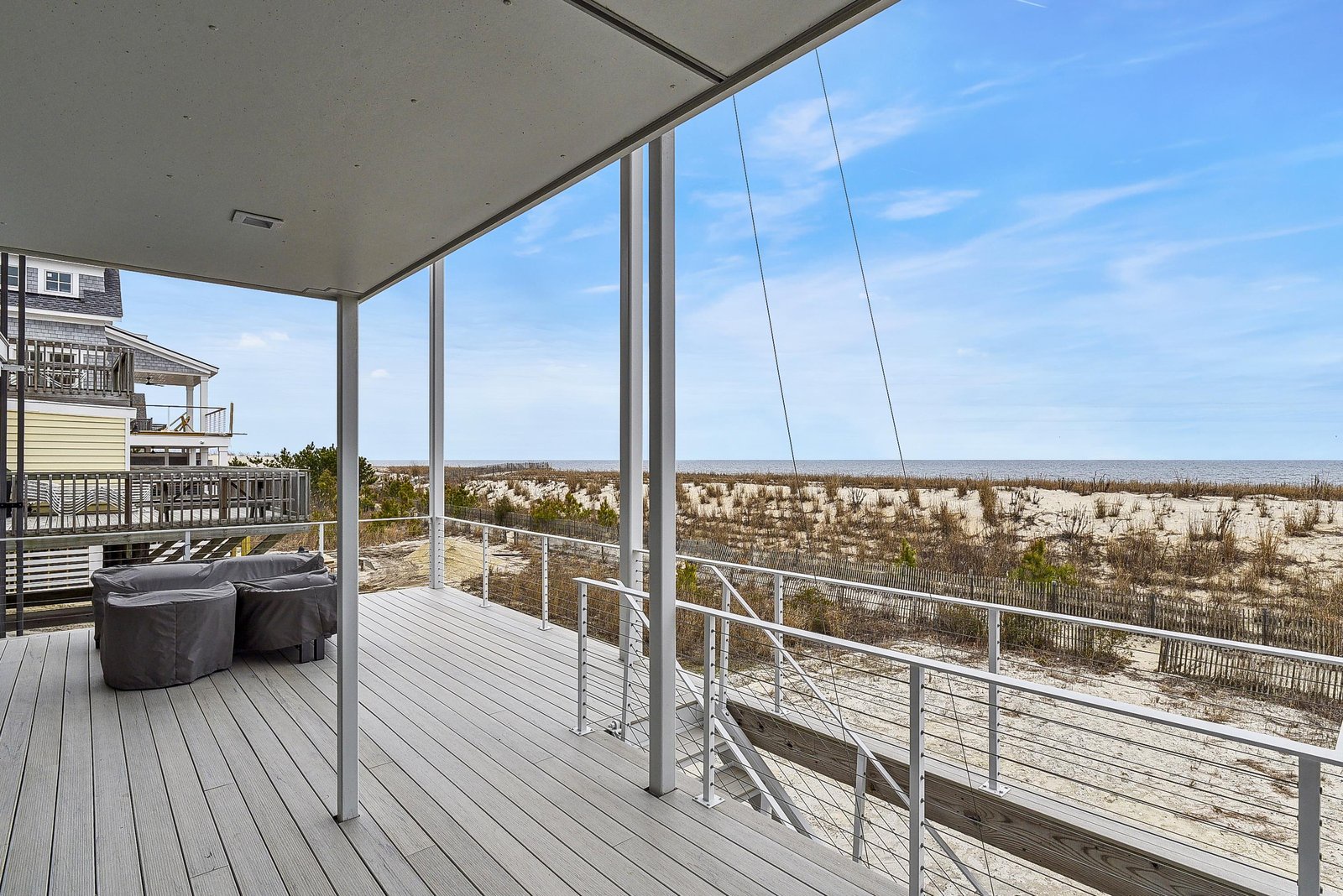 A covered deck with modern cable railing stretches toward sandy dunes and the sparkling ocean beyond—your front-row seat to coastal serenity.