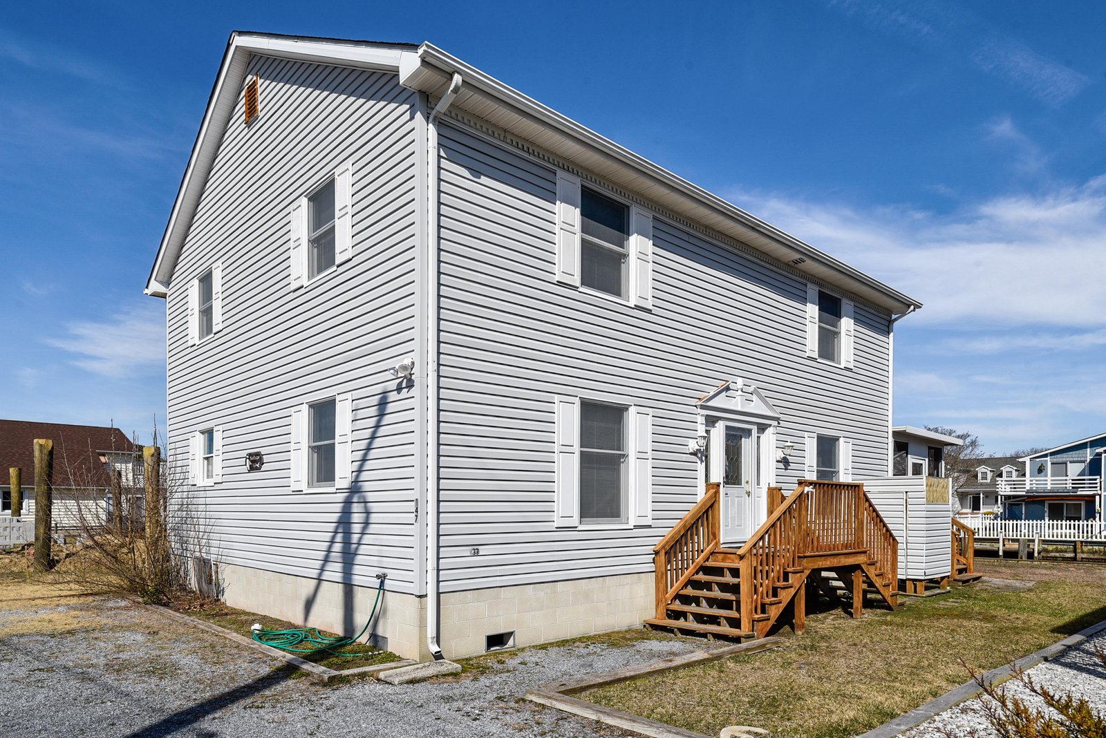 The property stands as a two-story home with welcoming wood deck entrance, set in a quiet neighborhood under clear blue skies.