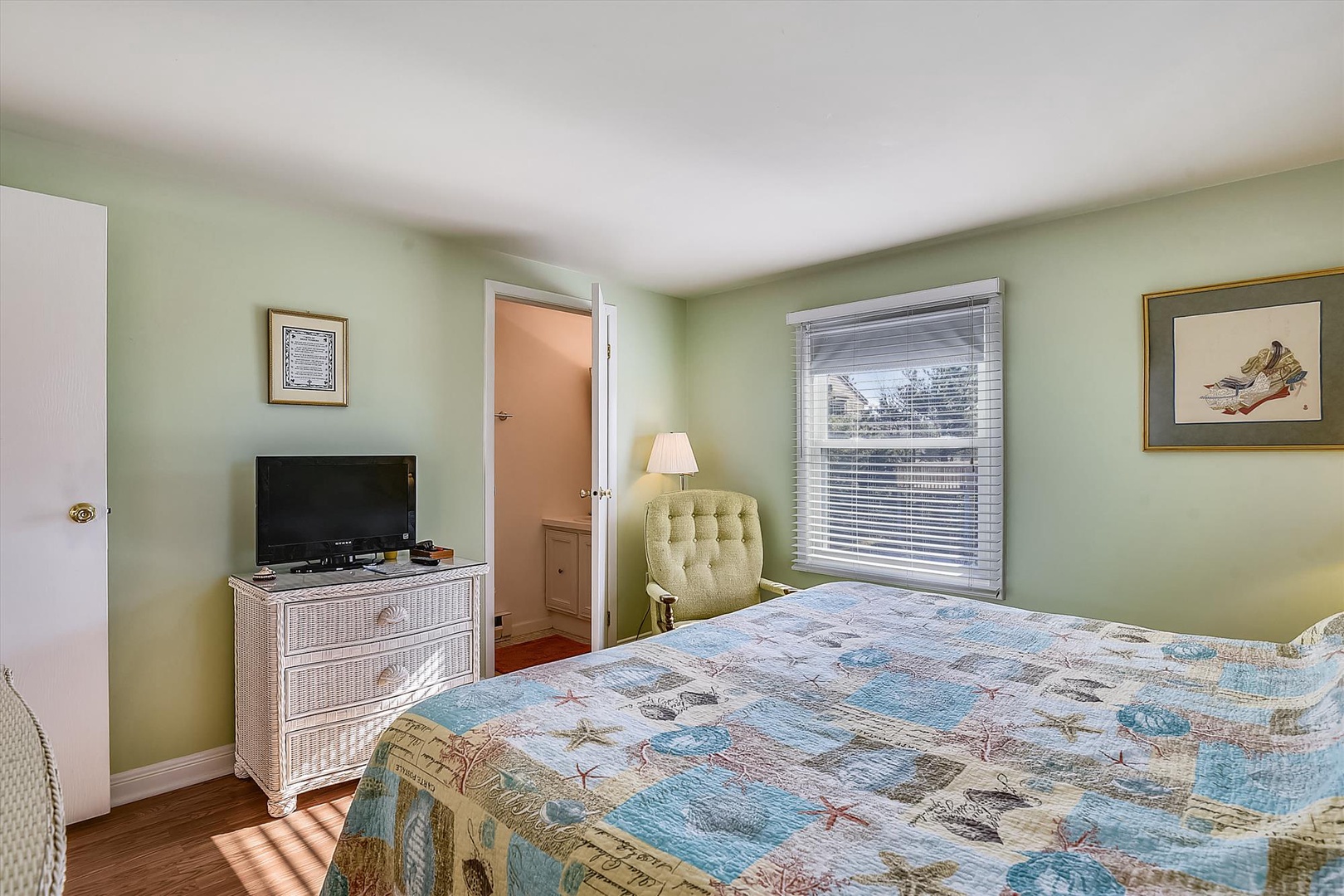 Settle into your cozy bedroom with calming sage-green walls, a plush quilt-topped bed, and natural light from the window. A TV and charming coastal artwork complete the restful retreat.
