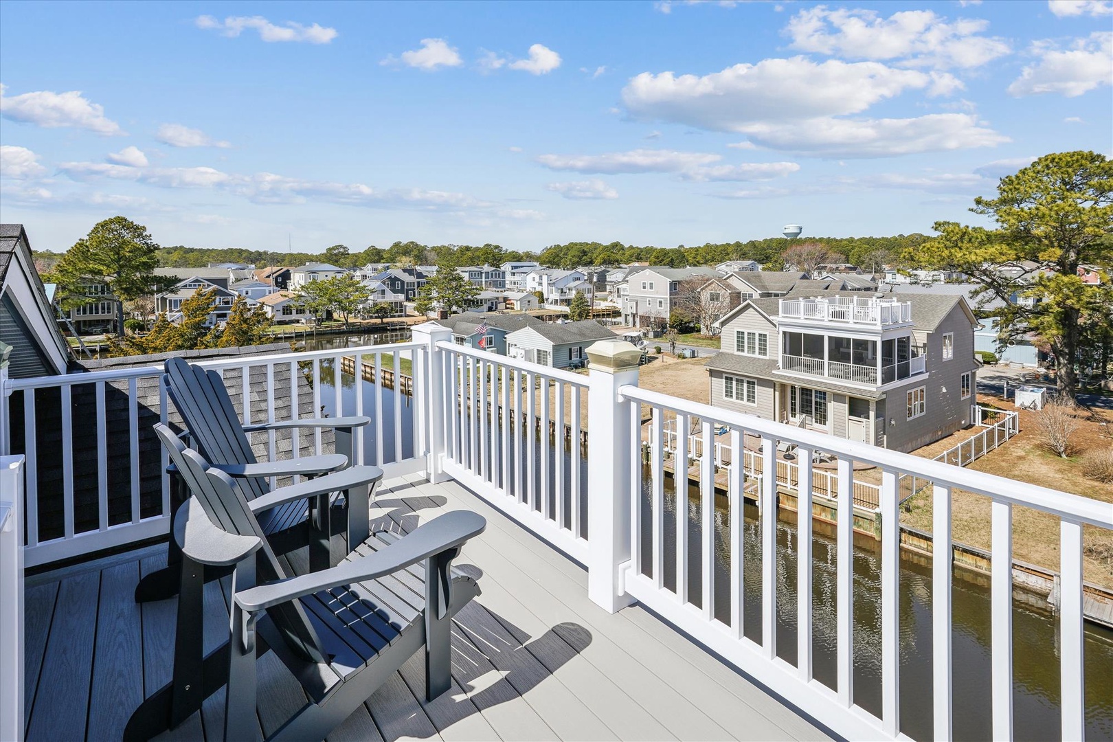 A bright balcony with classic Adirondack chairs where you can relax with morning coffee or evening drinks, overlooking a peaceful waterfront neighborhood framed by coastal pines.