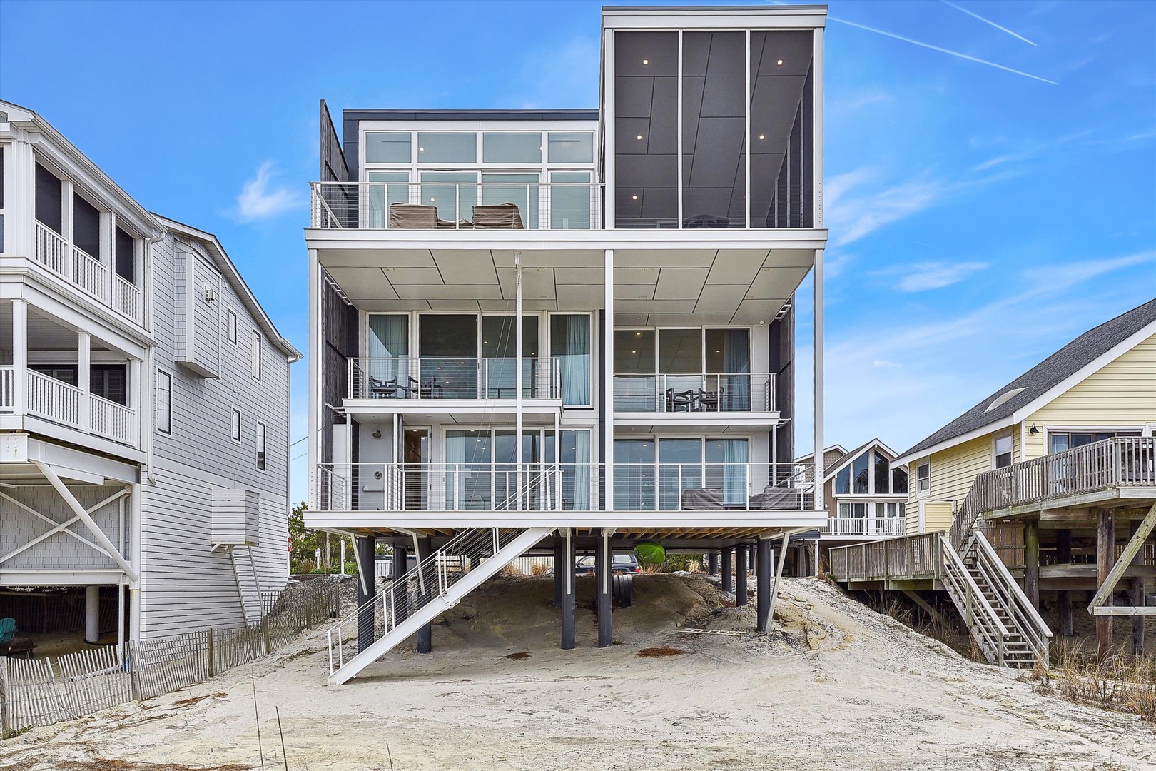 A modern, oceanfront beach house elevated on pilings with expansive glass walls and multiple balconies, situated in a coastal neighborhood with direct beach access.