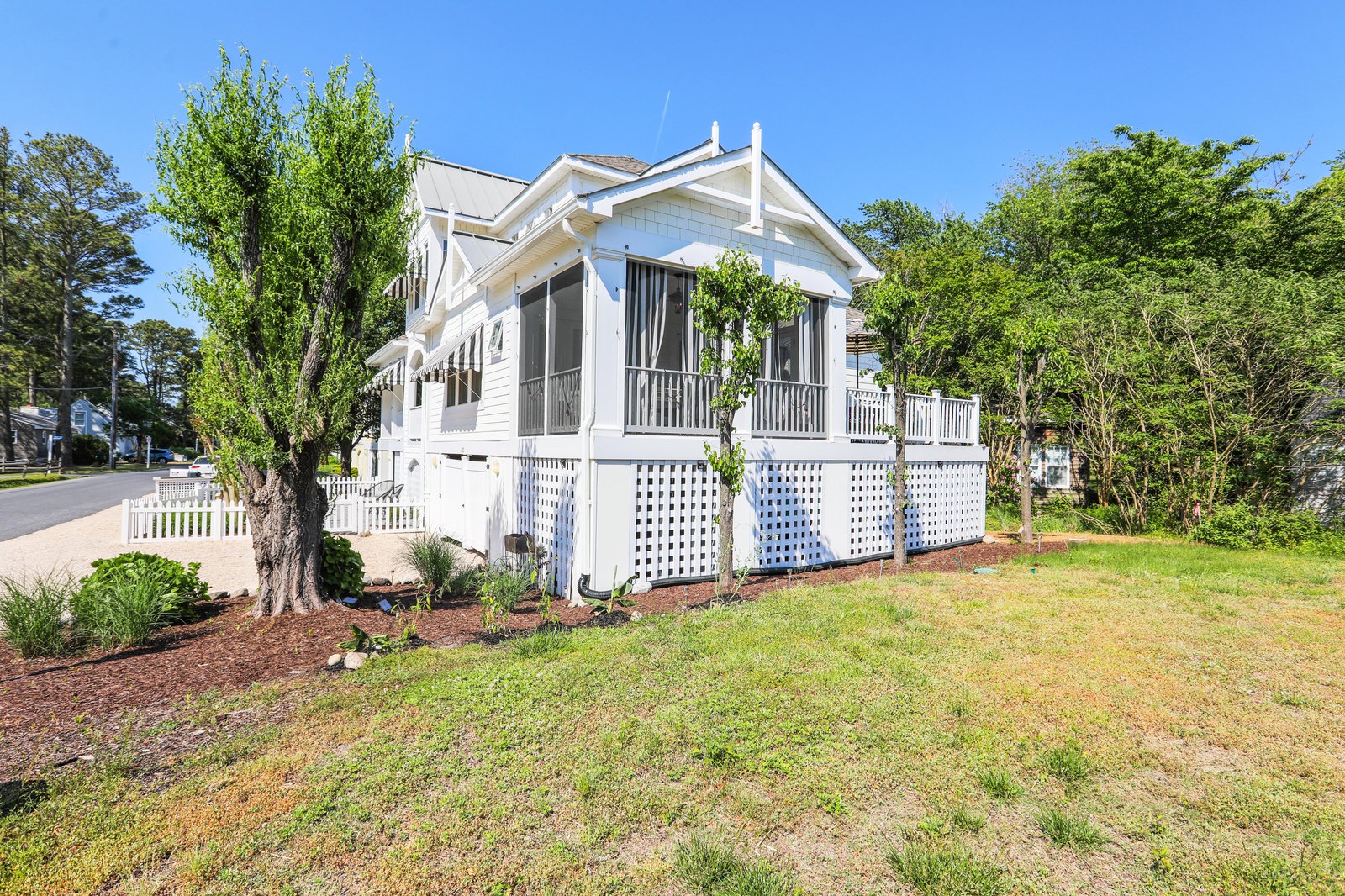 This charming white cottage with welcoming front porch sits nestled among mature trees in a peaceful residential setting, offering a classic coastal retreat feel.