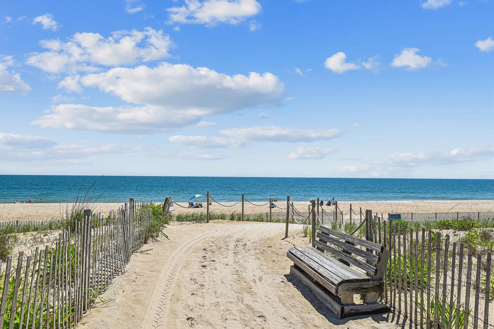 A sandy path leads through beach grass and weathered dunes to the turquoise waters beyond, where gentle waves meet pristine sand under an expansive coastal sky.