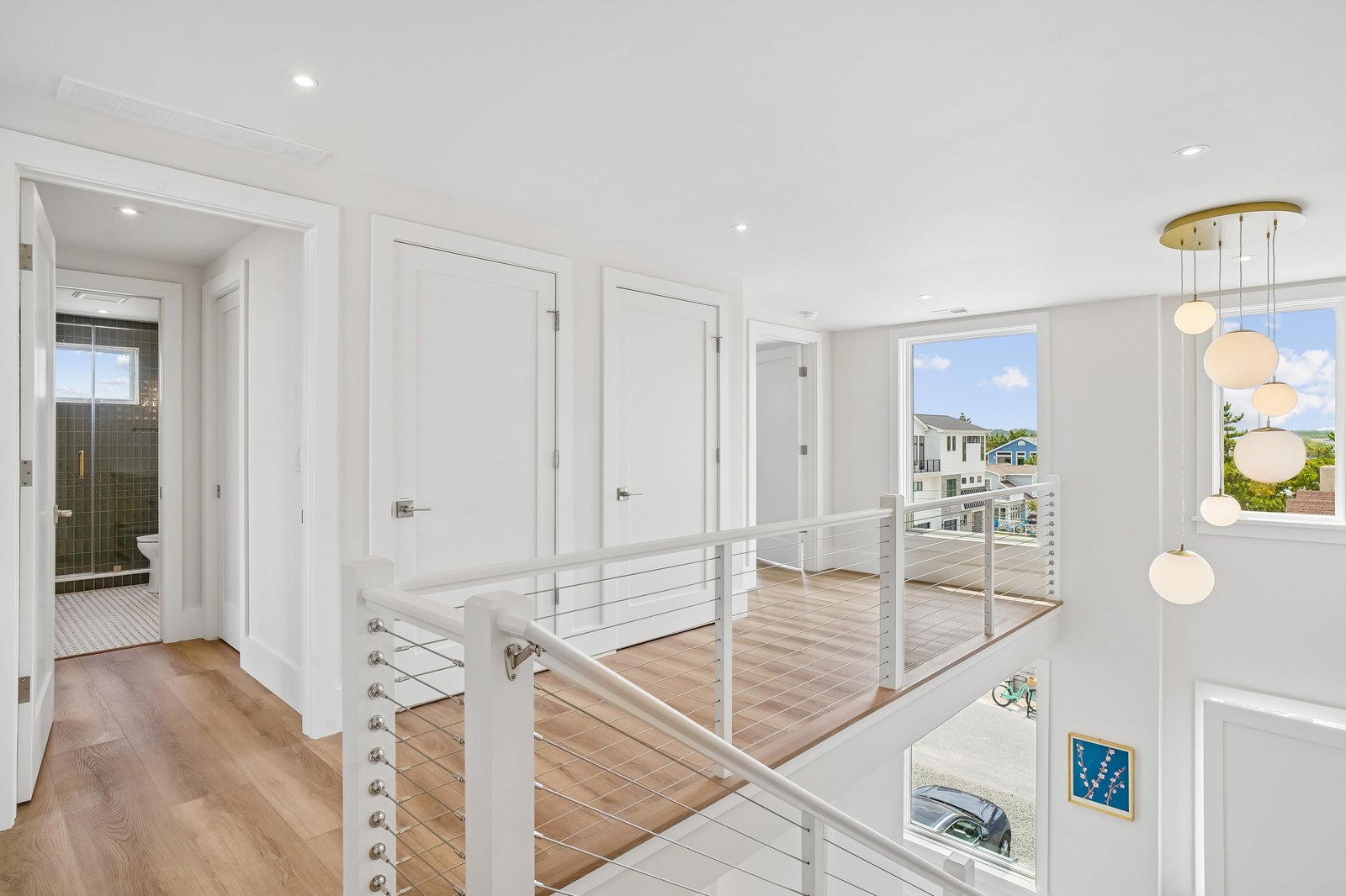 Light-filled modern hallway with warm wood floors, sleek railings, and bright windows framing views of the surrounding neighborhood and sky.