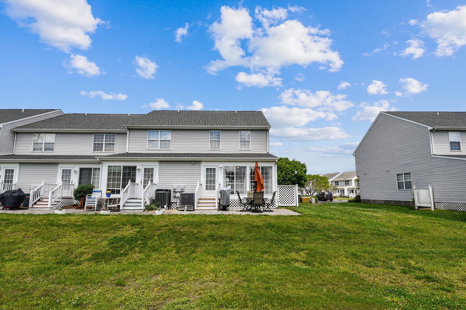Gray-sided townhome exterior with a spacious green lawn, white-fenced patios, and outdoor seating areas under open blue skies.