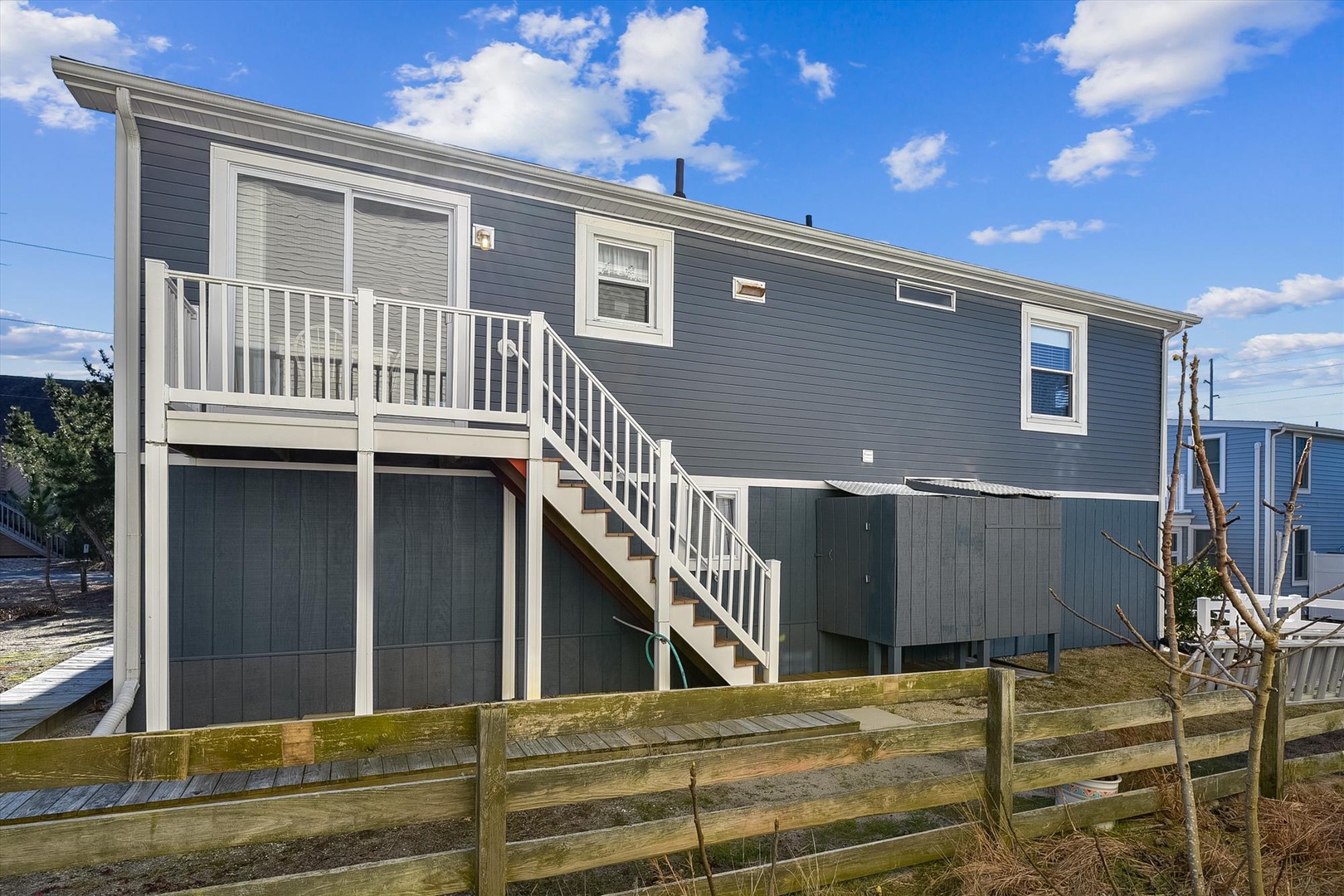 Modern coastal home with exterior stairs leading to elevated entrance, situated in a waterfront neighborhood beneath bright blue skies.