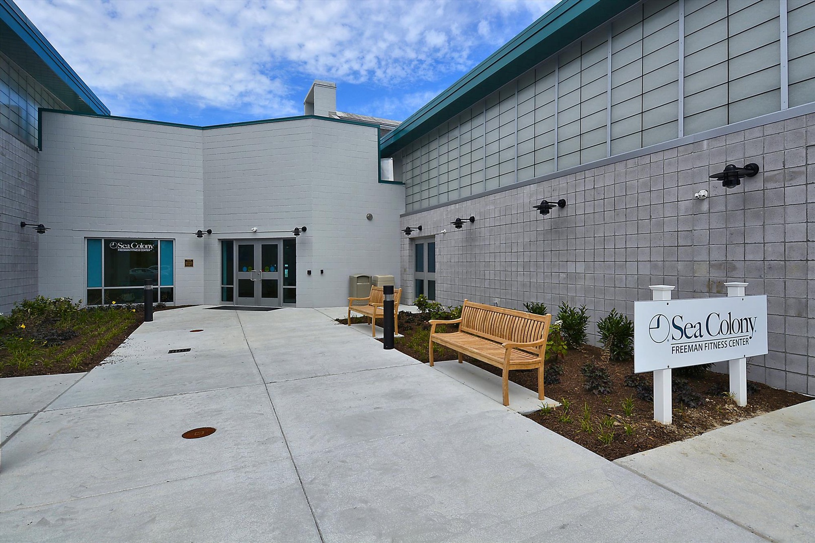 This modern building features clean lines and a welcoming courtyard entrance with benches and landscaped greenery under a bright blue sky.