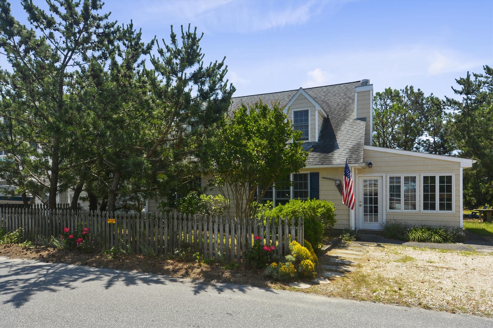 Classic coastal cottage set in a tranquil, tree-lined neighborhood with charming picket fence and American flag, welcoming guests to a peaceful vacation retreat.