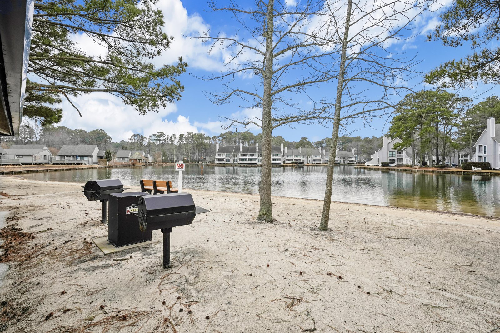 Lakefront community with BBQ facilities and tranquil water views, surrounded by seasonal trees and neighboring vacation homes. Sandy shoreline and benches provide an inviting communal gathering spot for guests.