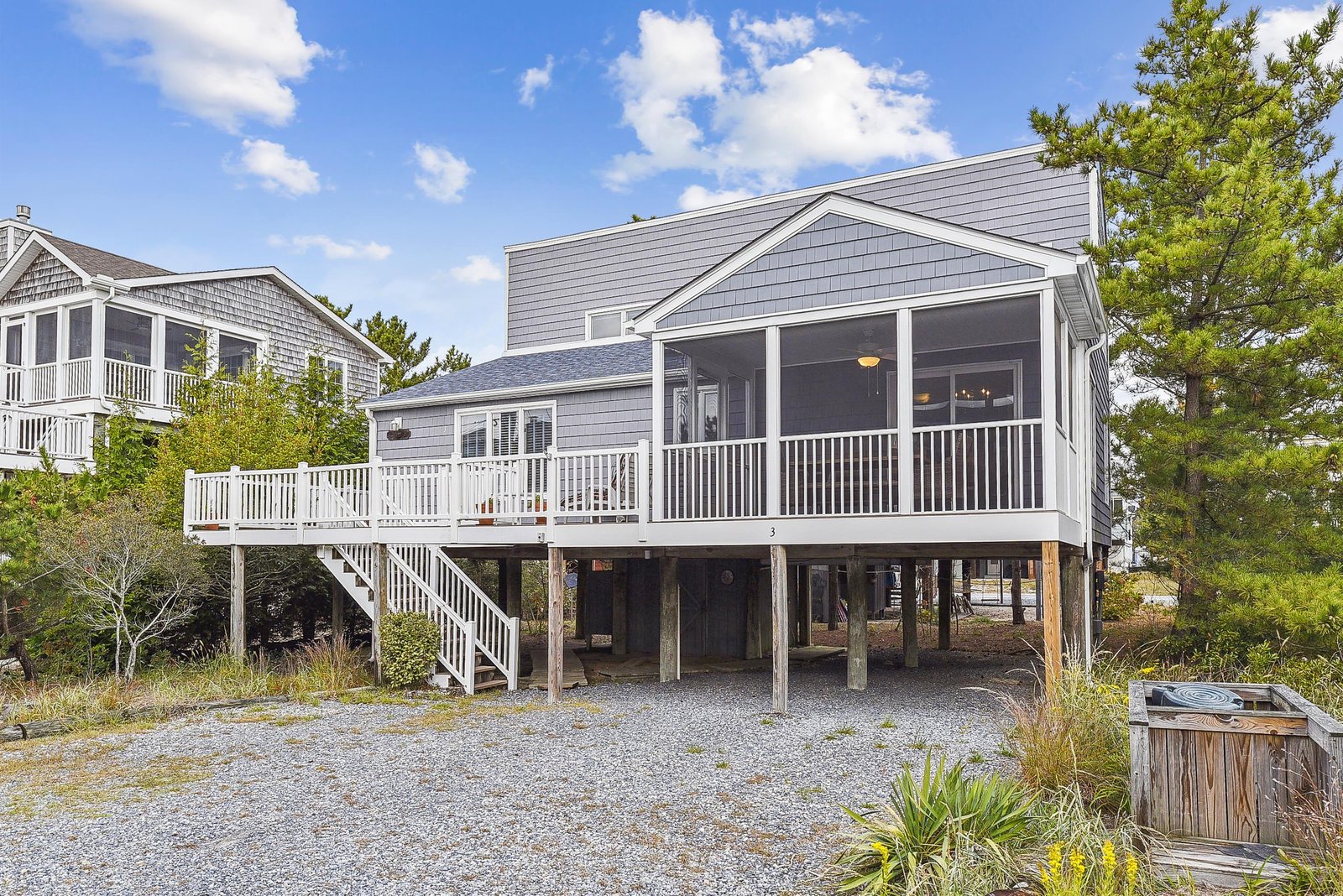 Coastal-style beach house featuring a screened porch, elevated deck, and gravel parking area, nestled in a quiet neighborhood near the shore.