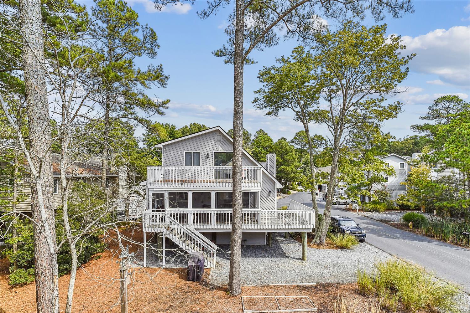 A two-story vacation home nestled among tall pines and mature trees, featuring spacious tiered decks and street-level parking in a peaceful wooded neighborhood.