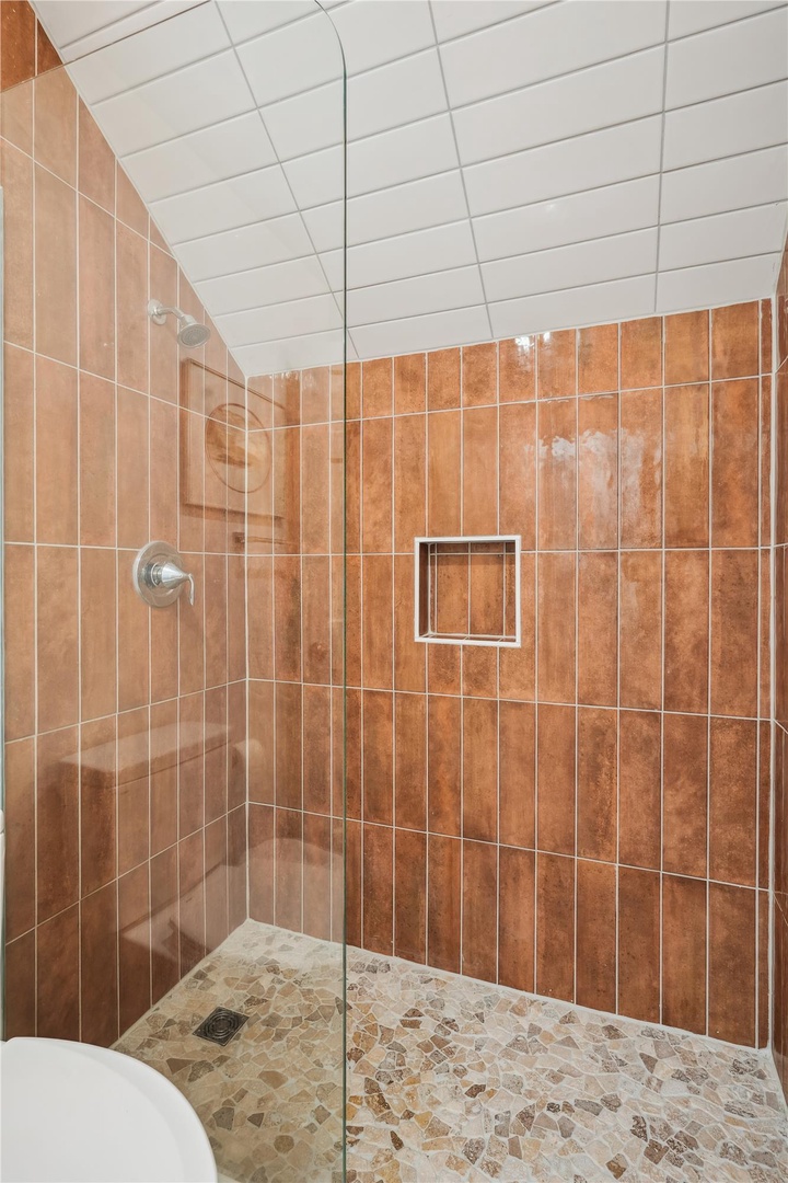 A spacious walk-in shower features warm terracotta tile, pebbled flooring for traction, a convenient built-in niche, and glass enclosure, creating a spa-like experience for your stay.