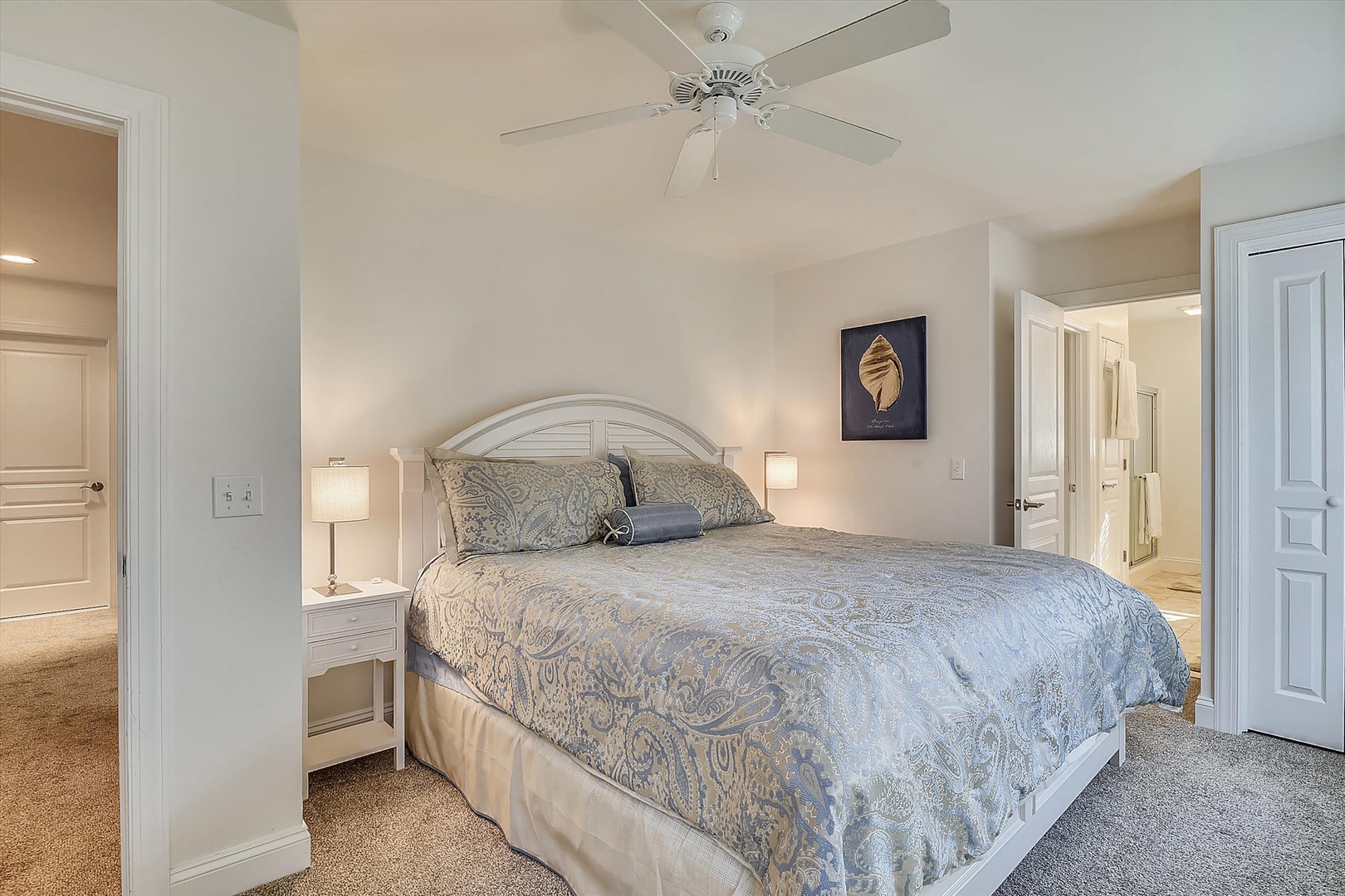 Drift off in a peaceful bedroom with plush bedding and soft lighting, where coastal art and a gentle ceiling fan set the scene for restful nights.