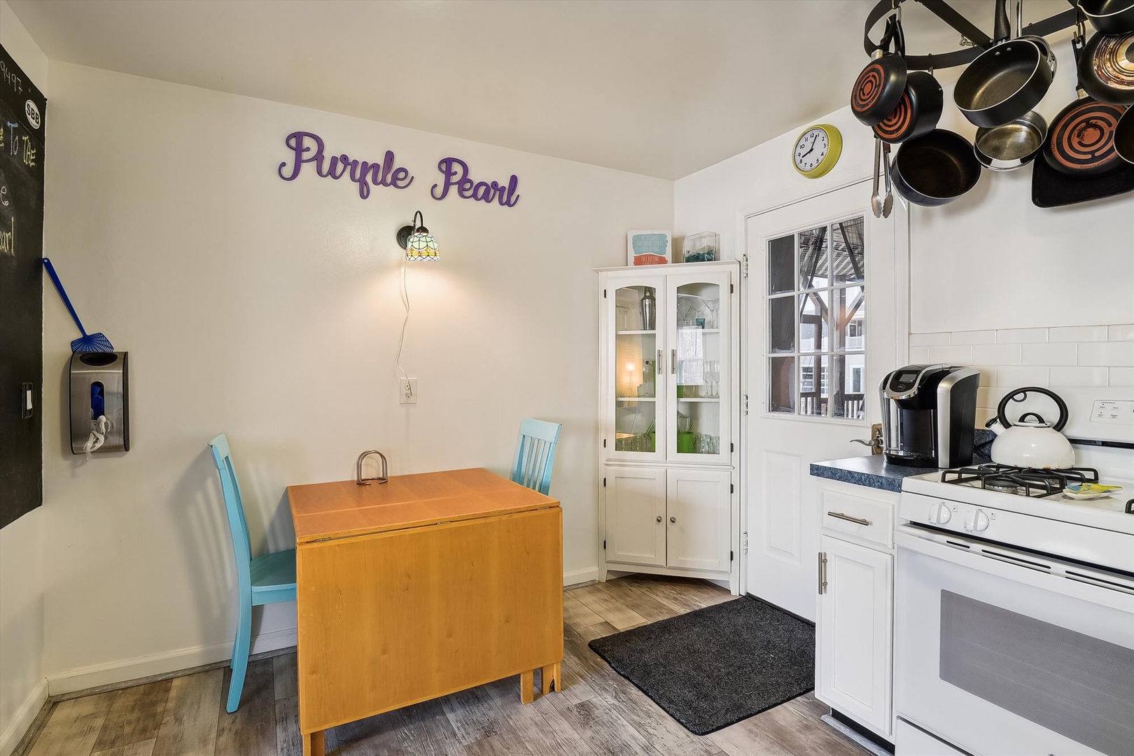 Your cozy kitchen features a compact dining area, charming purple signage, hanging cookware, and a gas stove with coffee maker ready for your morning brew.