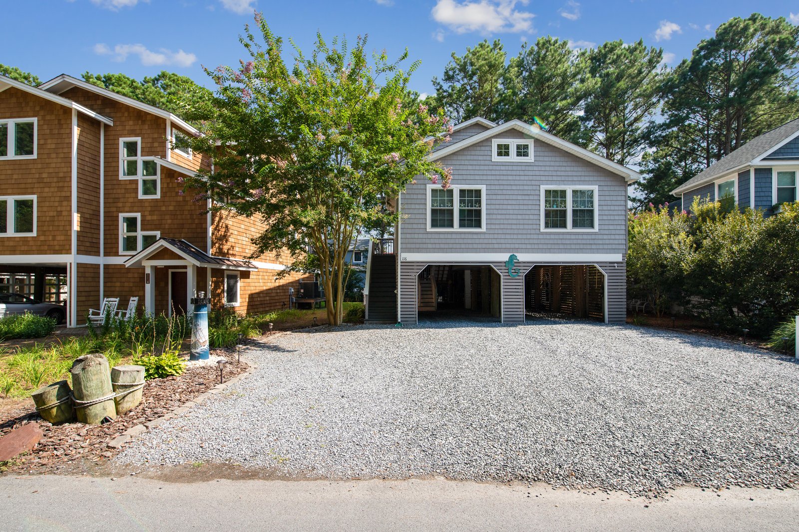 A coastal-style home with covered parking and a gravel drive, nestled in a quiet neighborhood surrounded by lush trees and neighboring properties.