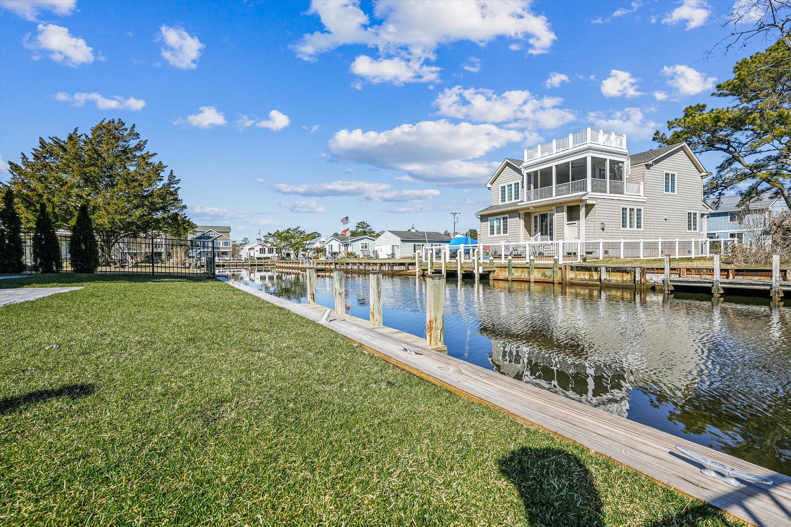 A spacious waterfront property featuring a private dock on calm waters, surrounded by lush green lawn and neighboring homes under a bright blue sky.