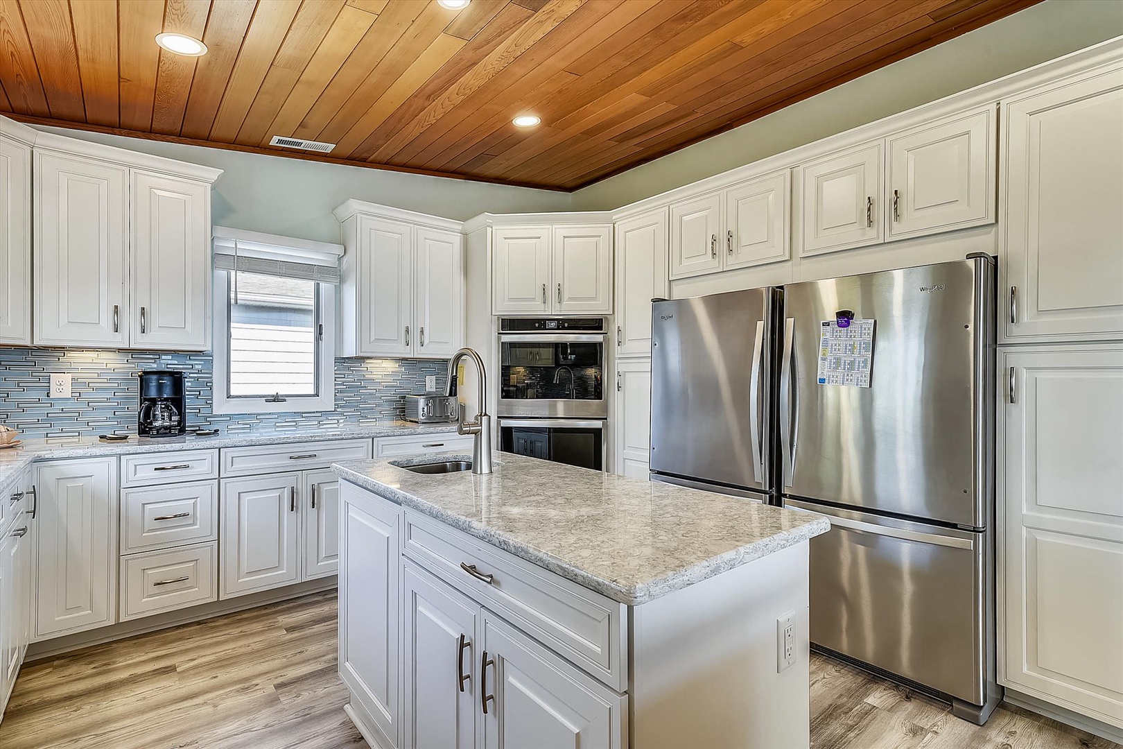 Your fully equipped kitchen features crisp white cabinetry, granite countertops, and stainless steel appliances beneath a warm wood-planked ceiling—ready for cooking meals and morning coffee together.