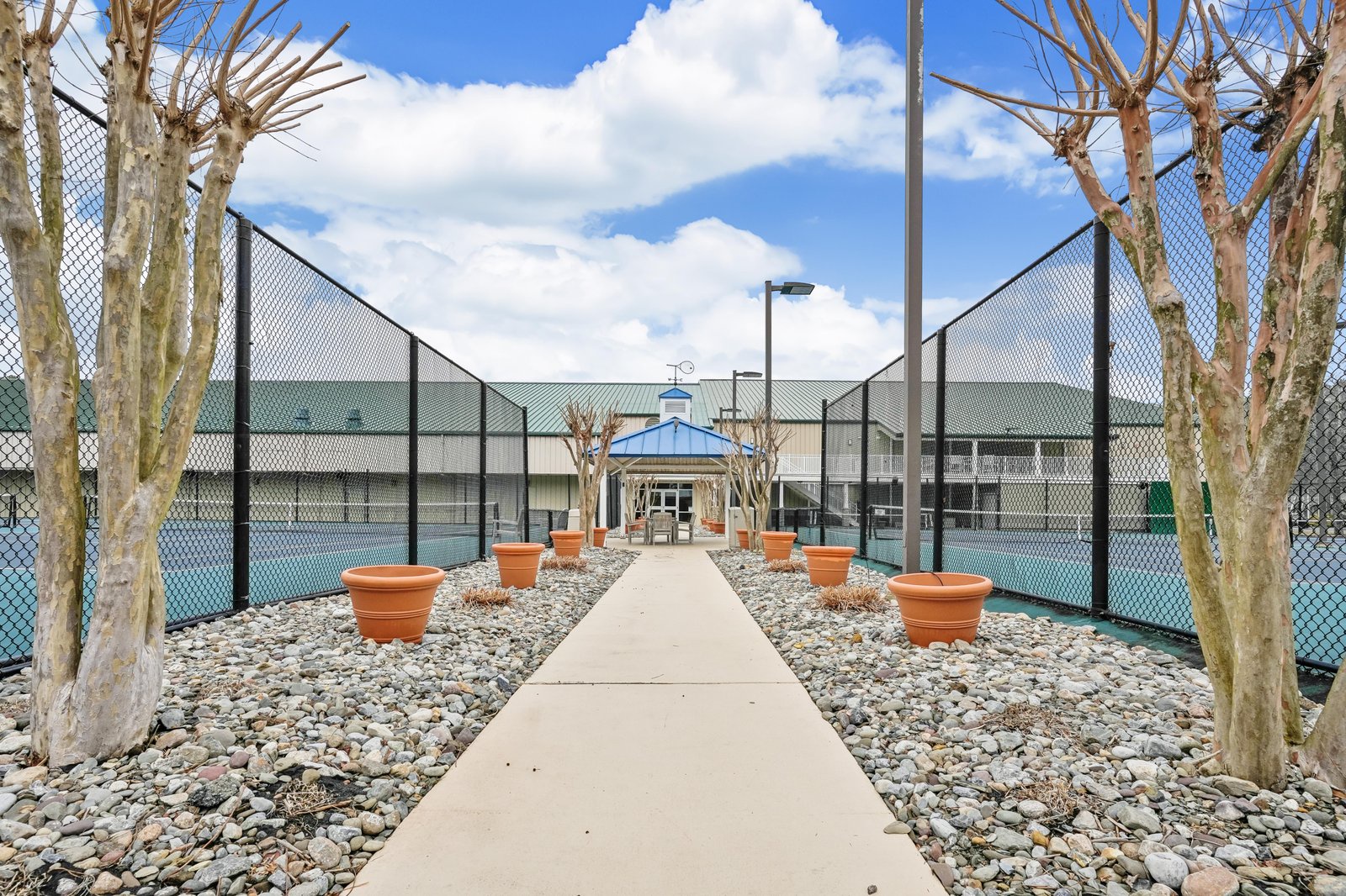 Welcoming entrance framed by palm trees and decorative planters, leading to the main building with tennis courts on either side under a bright blue sky.