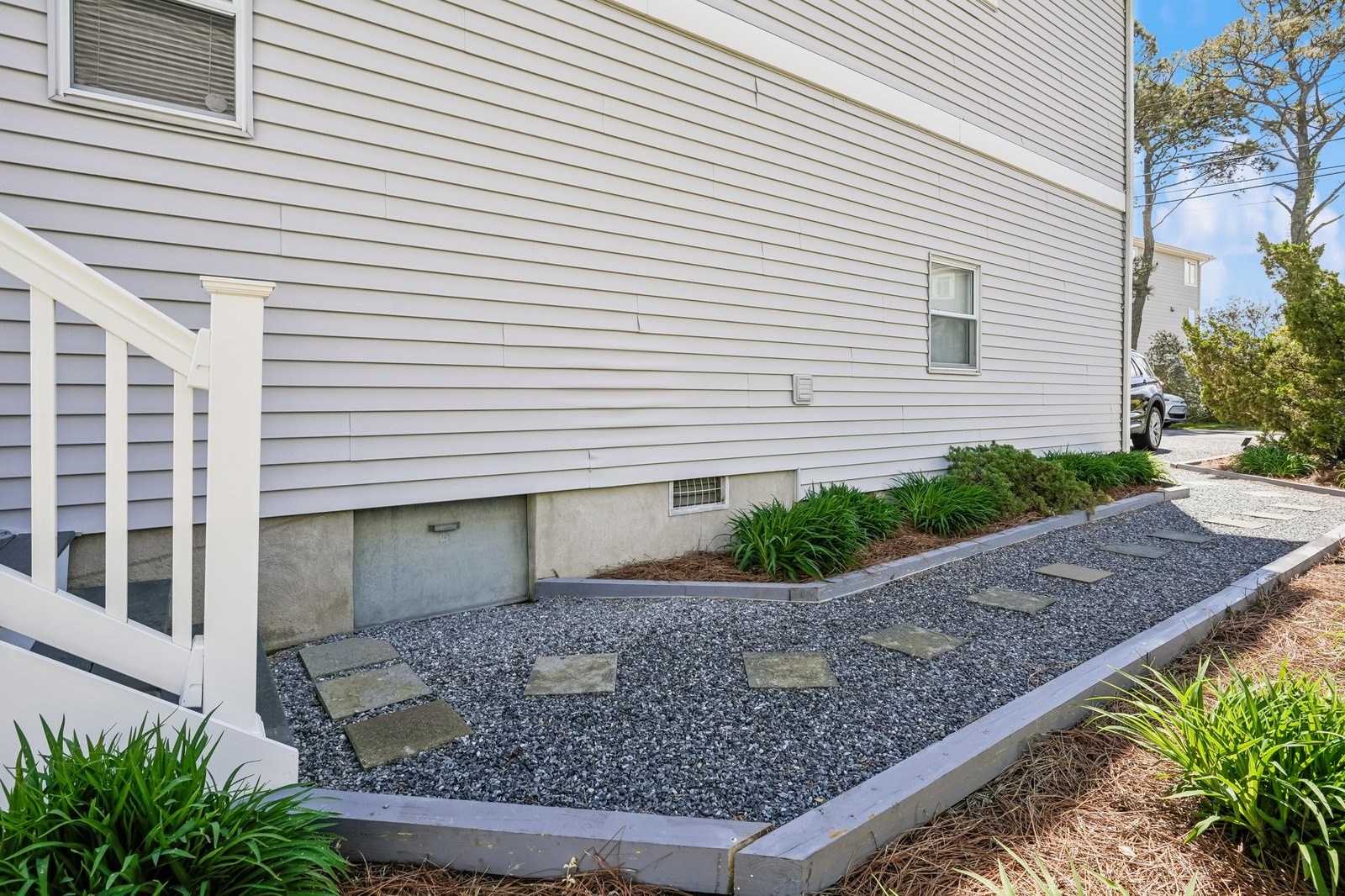 A tranquil gravel pathway lined with stepping stones leads along the side of this coastal property, framed by greenery and glimpses of blue sky—a peaceful arrival path to your beachside retreat.