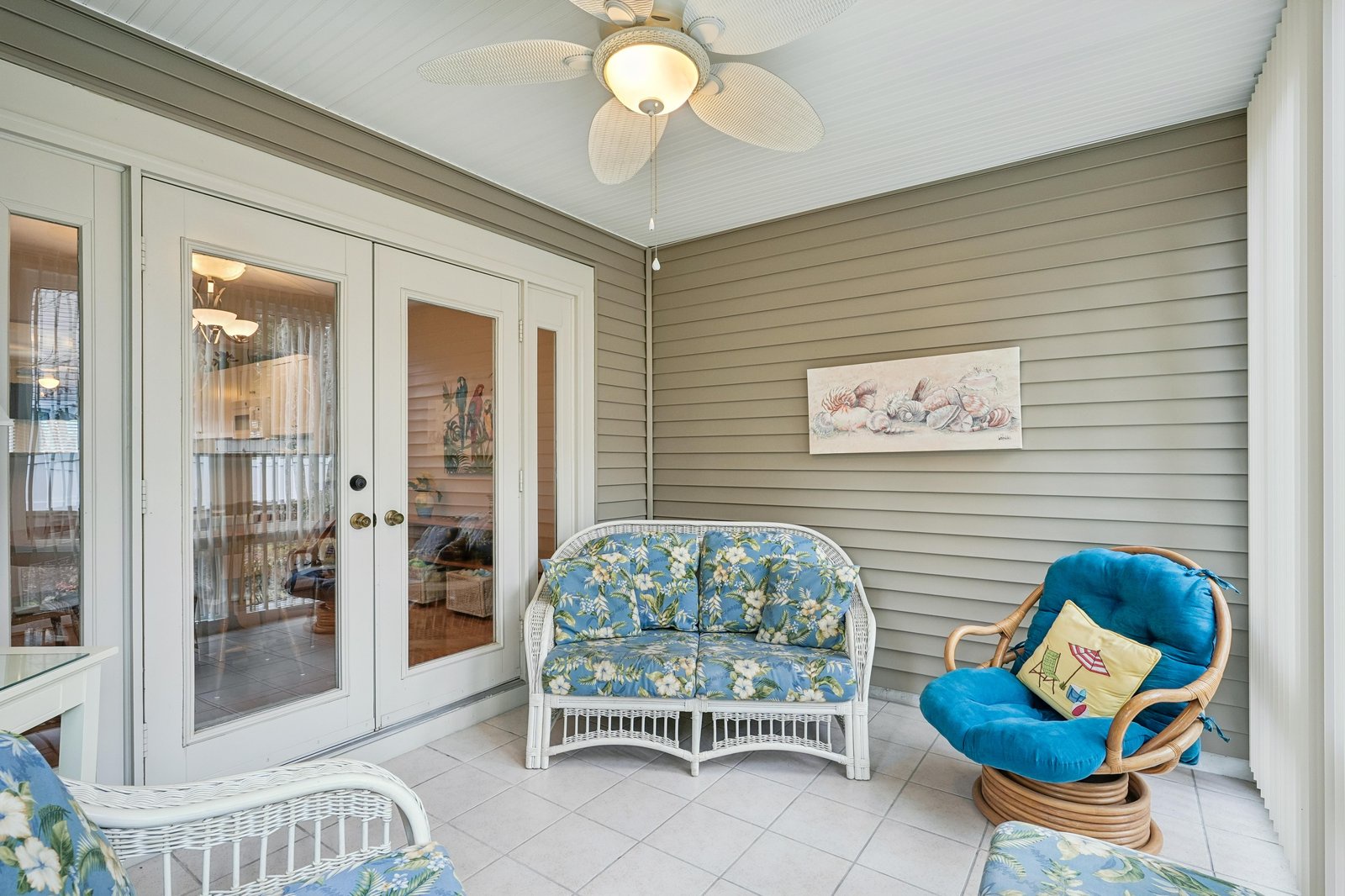 An enclosed patio offers a peaceful retreat with coastal decor. Ceiling fan above, comfortable seating, and glass doors welcome you to unwind in this breezy, private space.